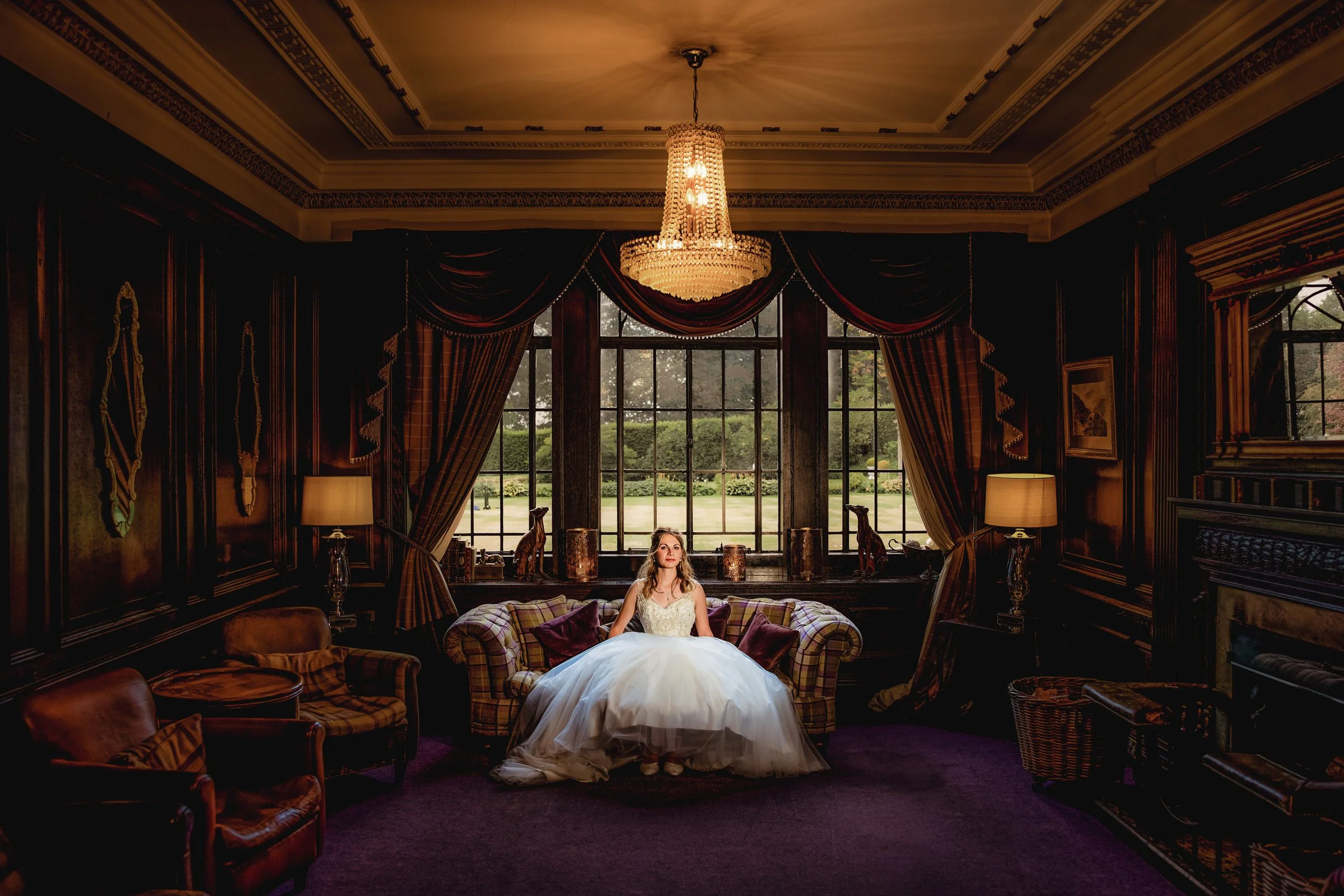 A woman in a wedding dress sitting on a sofa in a dark, richly decorated room with large window and curtains, chandelier hanging from ceiling, and lamps on either side of the window.