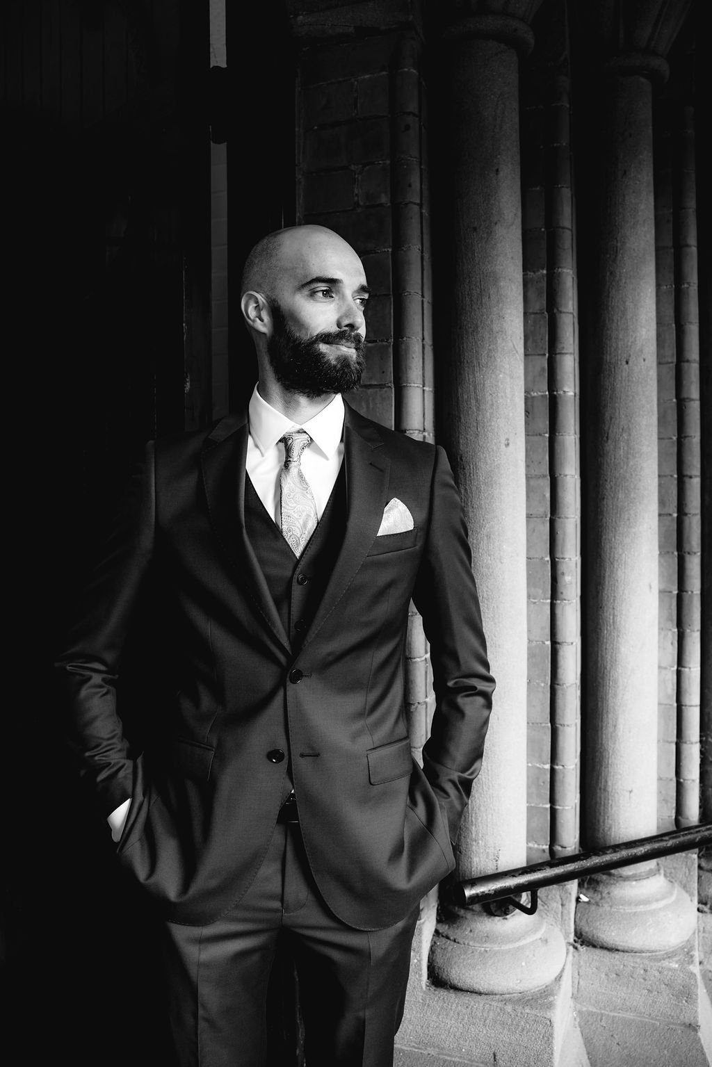 A man in a suit with a pocket square, standing outdoors near brick columns, looking to the side, in black and white.