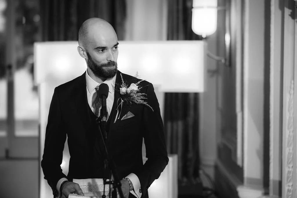A man in formal attire, with a beard and bald head, giving a speech at a microphone. He is wearing a dark suit with a boutonniere. The background shows a well-lit, elegant indoor setting.