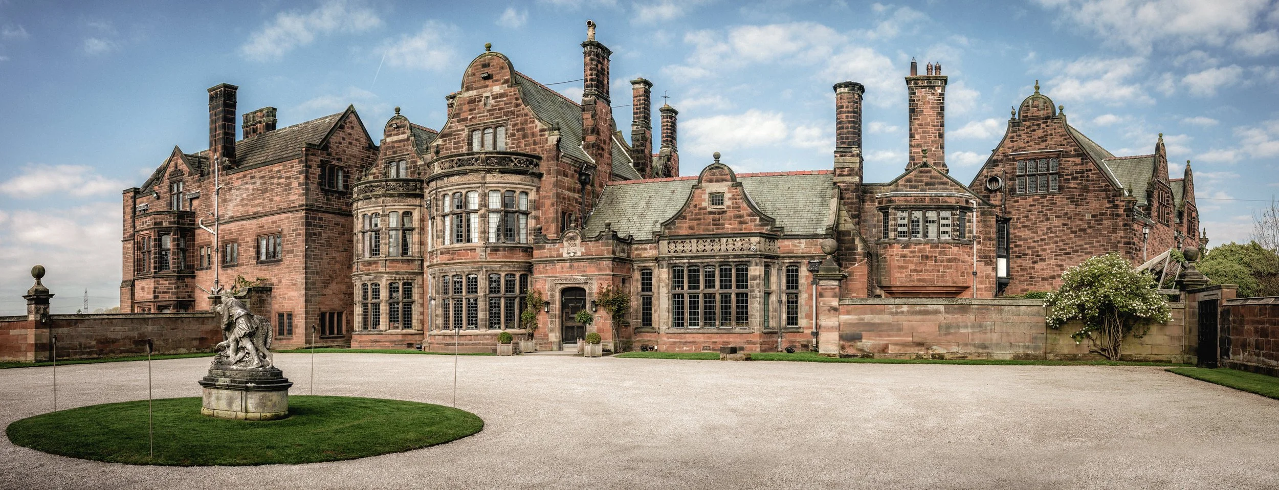 A large historic red-brick mansion with multiple chimneys and ornate windows, surrounded by a gravel courtyard, a statue of a horse and rider on a grassy island, and a stone wall with gates, under a partly cloudy sky.