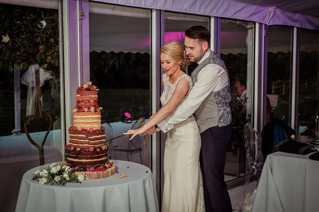 A bride and groom cutting a multi-tiered naked cake decorated with berries and flowers at a wedding reception.