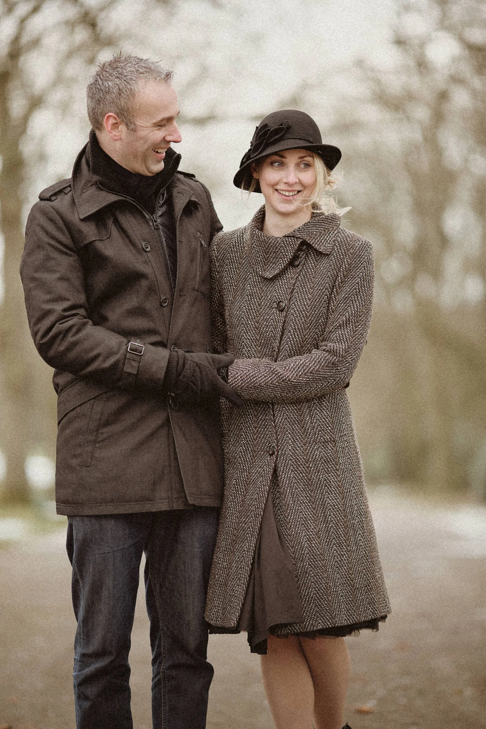 A man and a woman dressed warmly, smiling and walking outdoors in a park with leafless trees.