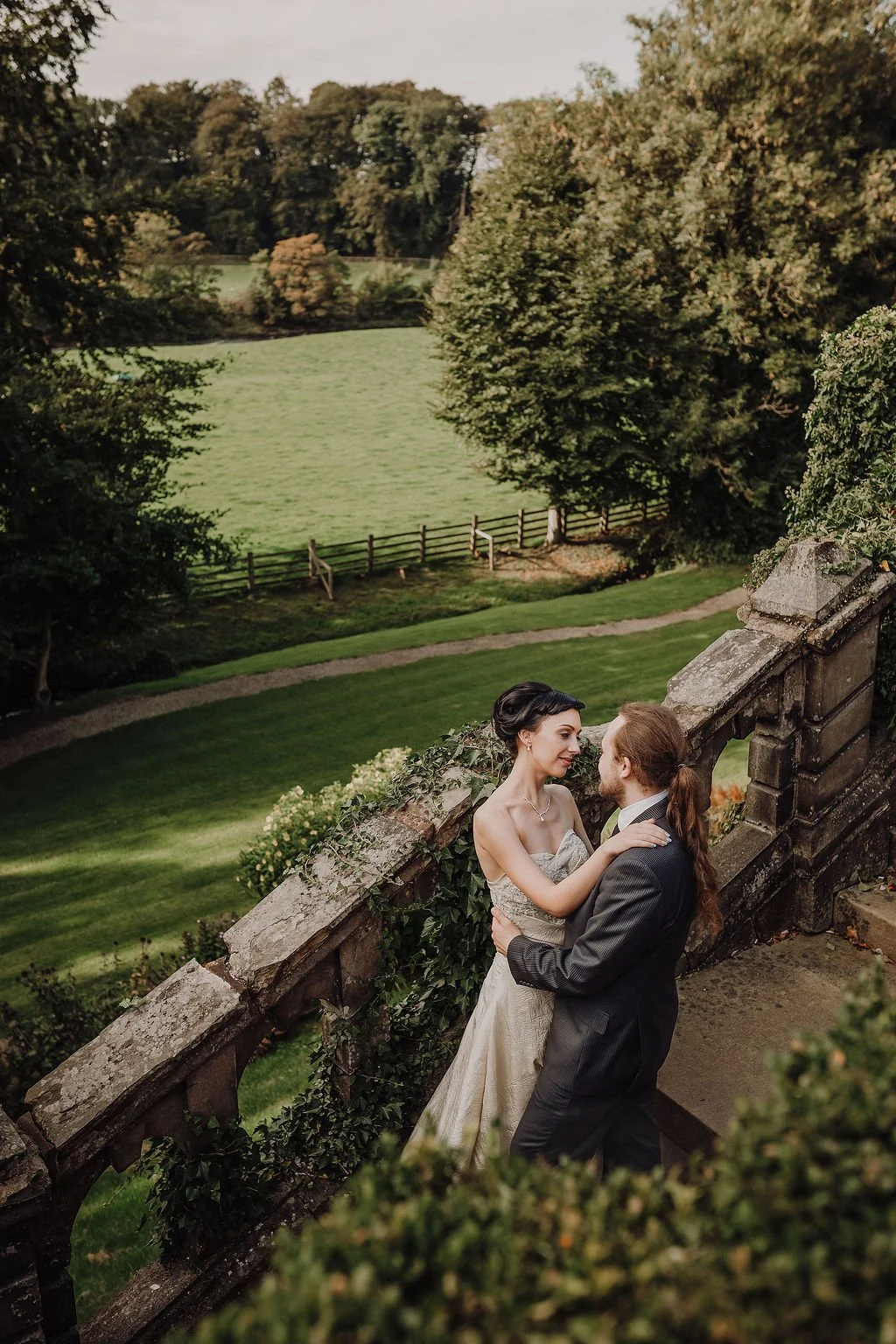 A bride and groom standing close together on a stone staircase outdoors, surrounded by greenery, looking into each other's eyes.