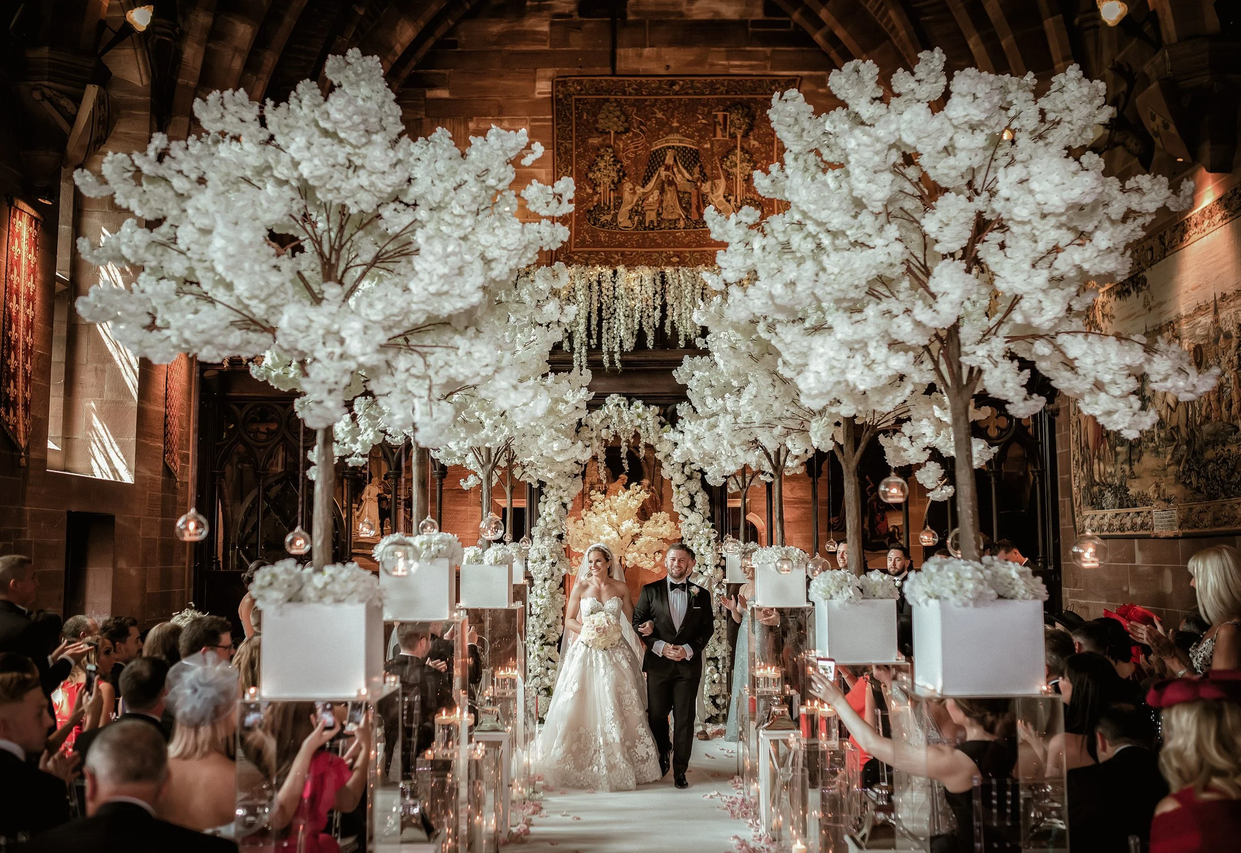 A bride and groom walking down the aisle at a wedding ceremony inside a church decorated with large white flowering trees and floral arrangements. Guests are seated on both sides, some taking photos.