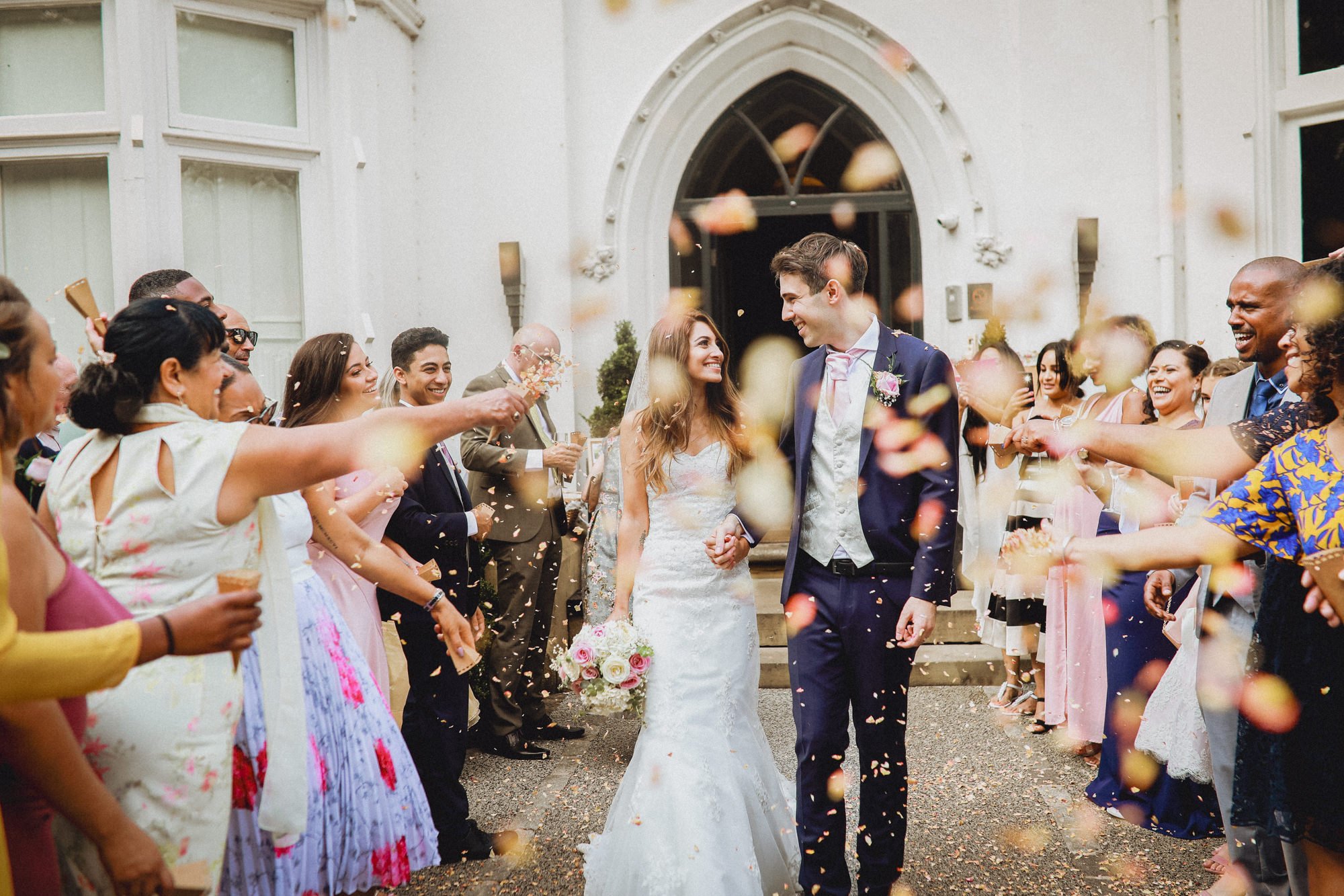 A bride and groom smiling and holding hands outside a wedding venue, surrounded by guests throwing flower petals.