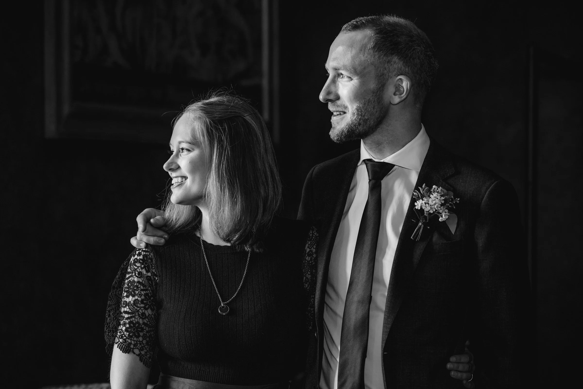 A black-and-white photo of a smiling woman and a man in formal attire, standing close together. The woman has shoulder-length hair and is wearing a lace-sleeved top with a necklace. The man has short hair, a beard, and is wearing a suit with a bouton