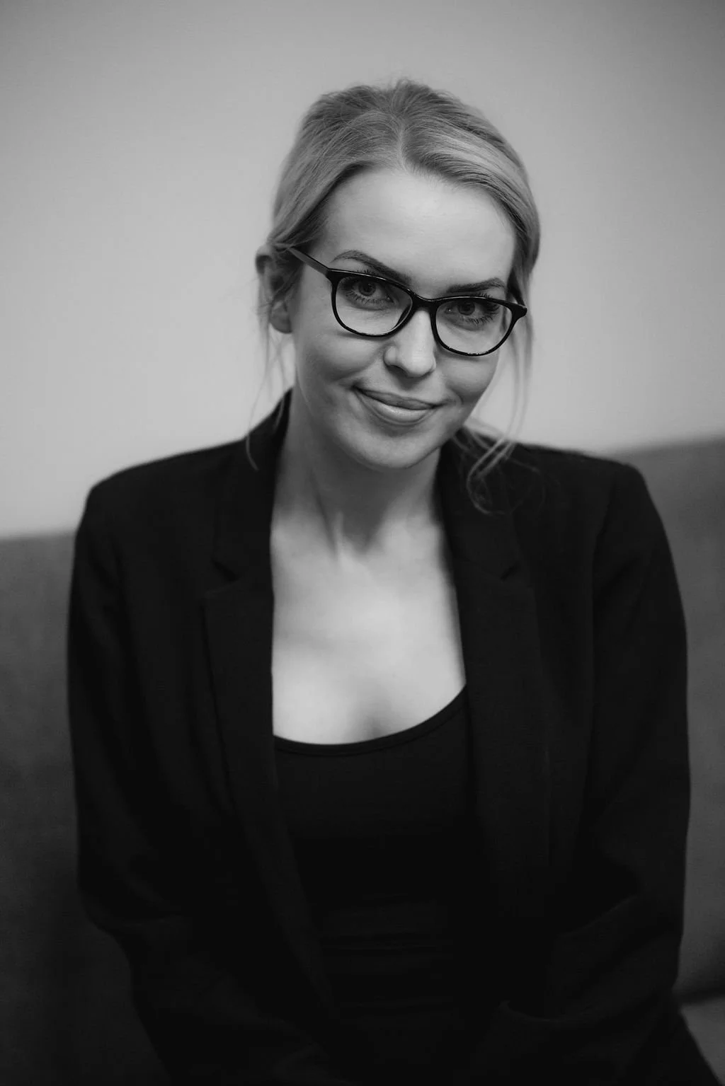 A black and white portrait of a woman with blonde hair and glasses, wearing a black blazer and shirt, smiling slightly at the camera.