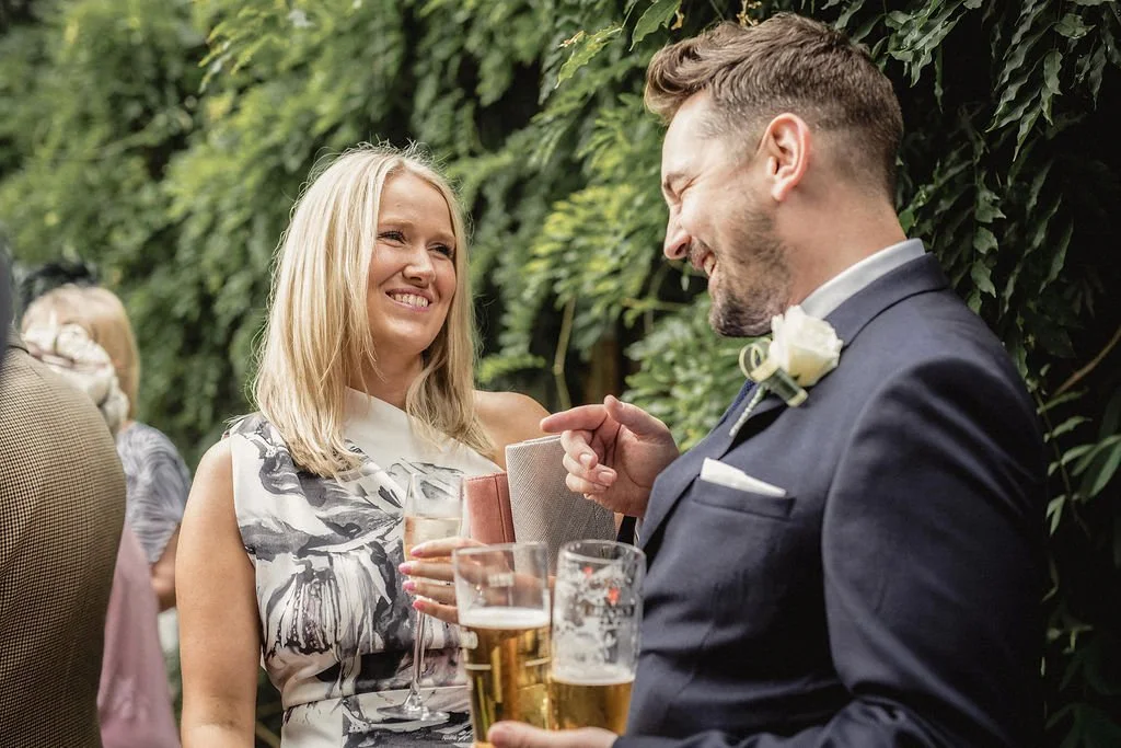 A man and woman at a social event, smiling at each other, with drinks in hand, dressed in formal attire, standing outdoors against a green leafy background.