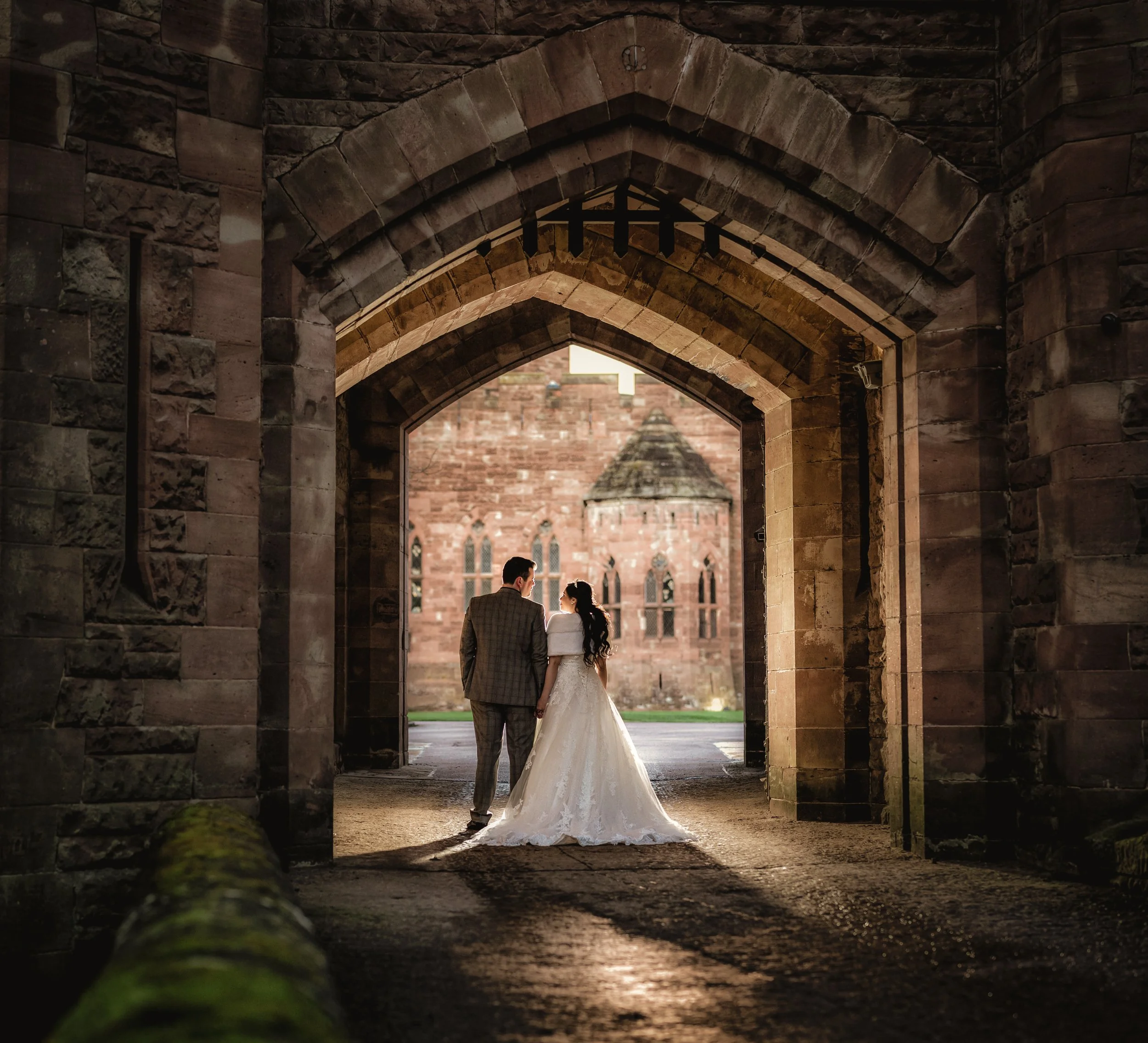 A bride and groom standing hand in hand in a stone archway, with a church in the background.