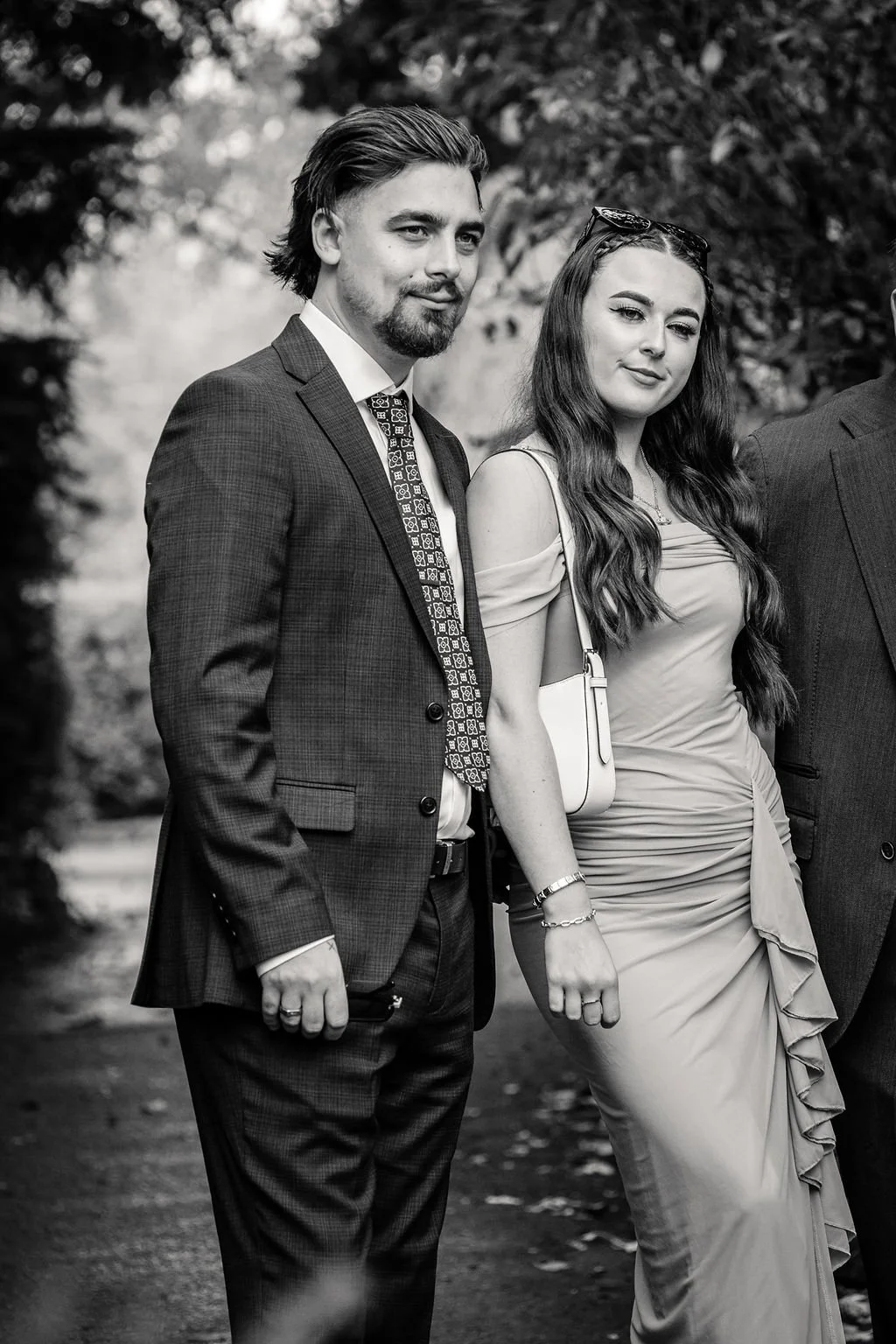 A black and white photo of a young man and woman standing outdoors, dressed in formal attire, with trees in the background.