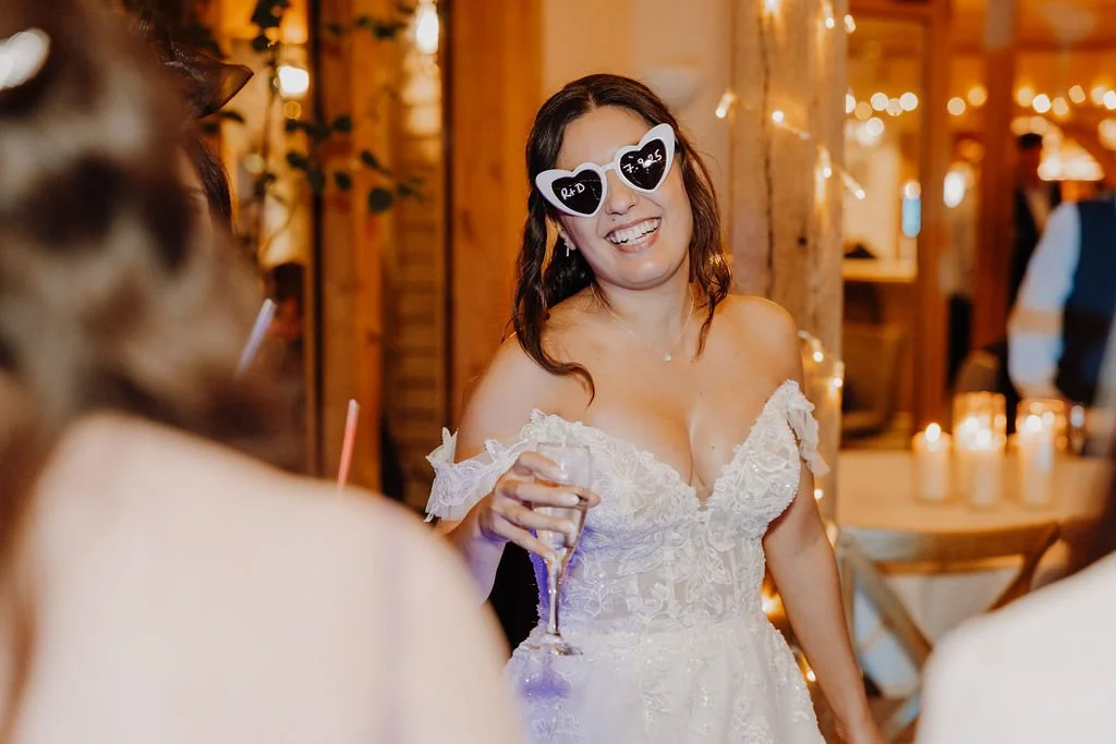 A woman in a wedding dress with heart-shaped sunglasses at a wedding reception, smiling and holding a glass of champagne.