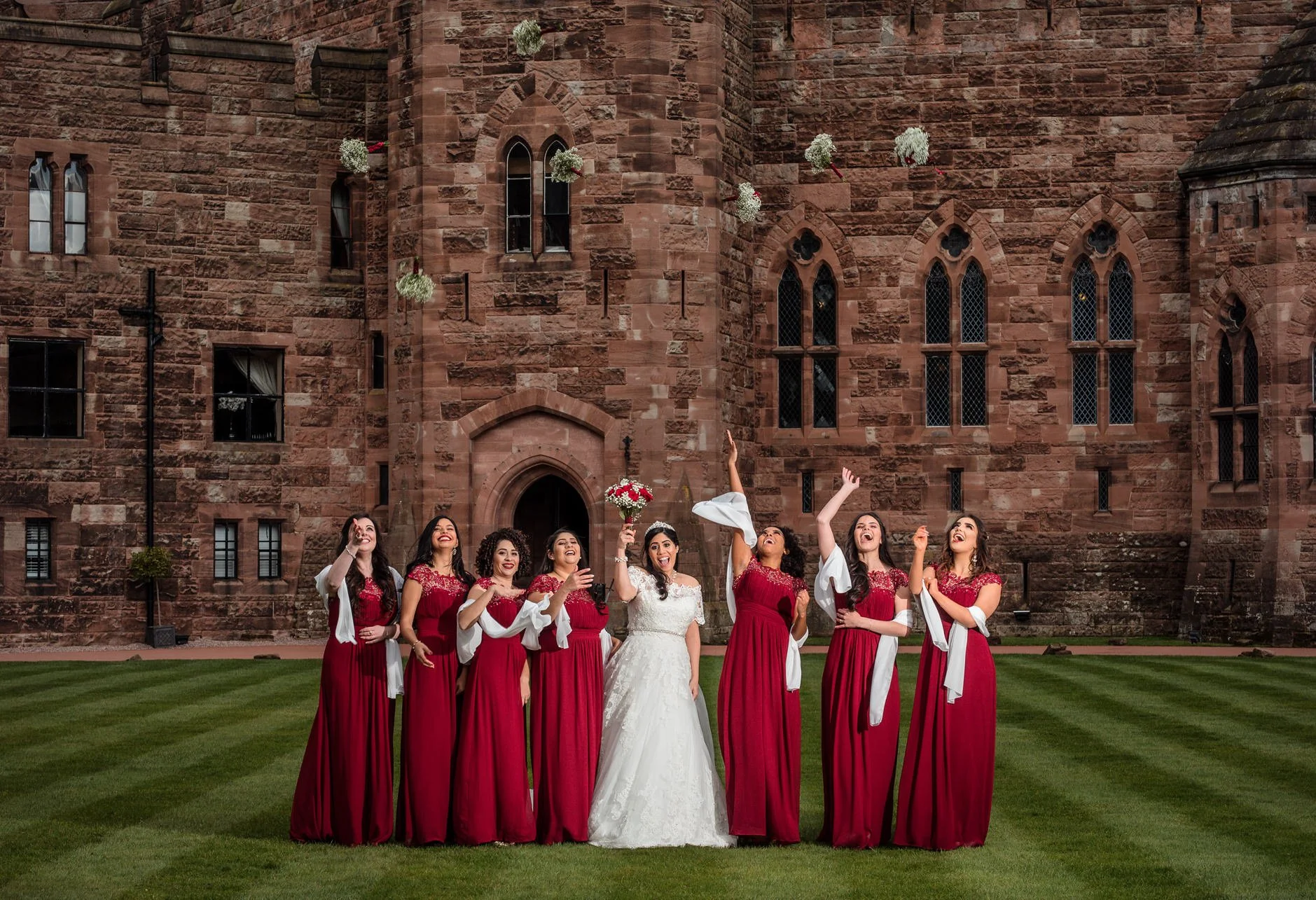 Bride in white wedding dress and seven bridesmaids wearing red dresses toss their bouquets into the air standing on a lawn in front of a large stone castle.