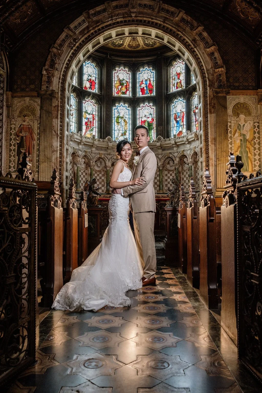 A bride and groom stand together inside a church with stained glass windows and wooden pews. The bride wears a white wedding gown and the groom wears a beige suit. They are smiling and looking at the camera.