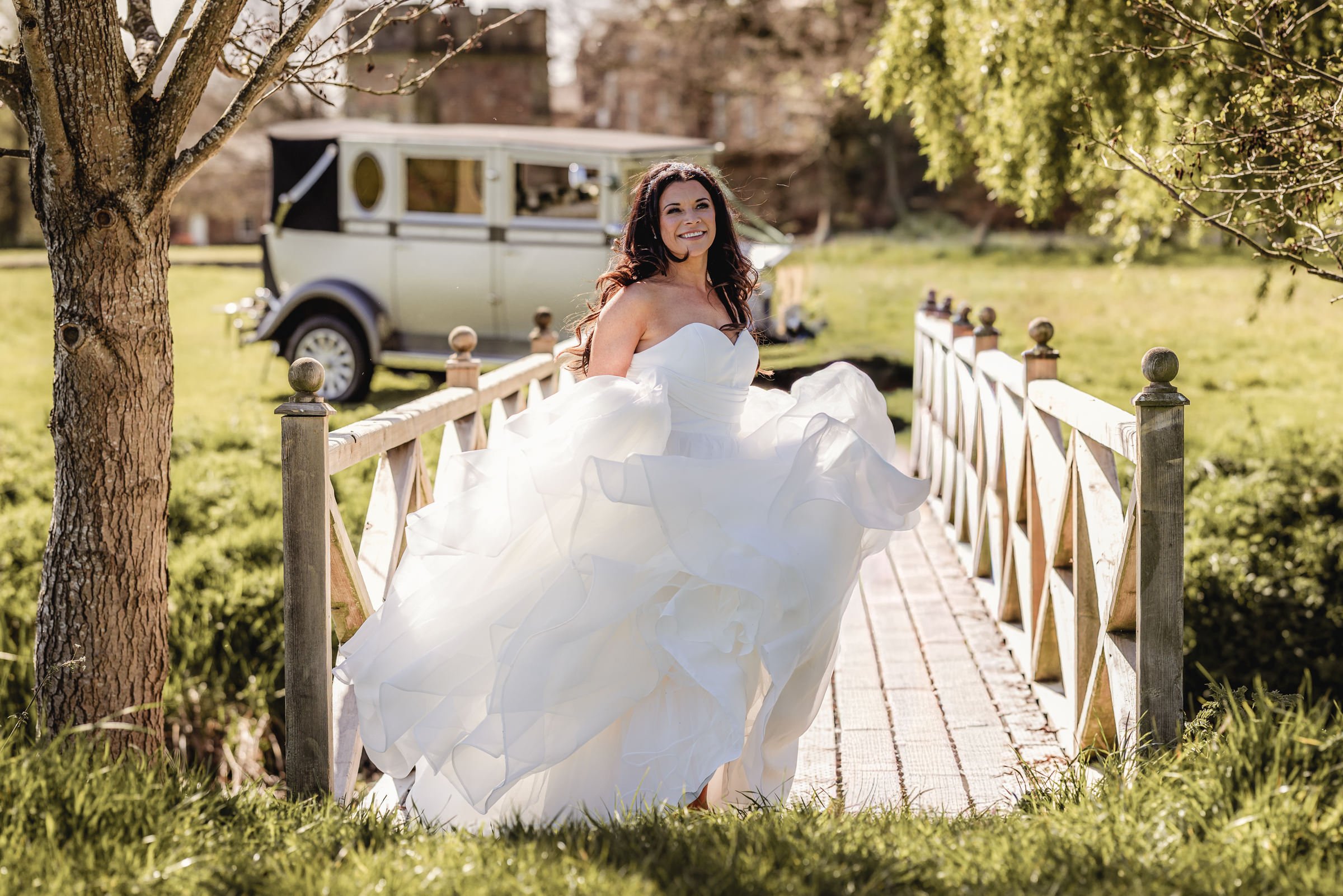 Bride in a white wedding gown walking on a small wooden bridge outdoors with a vintage car in the background