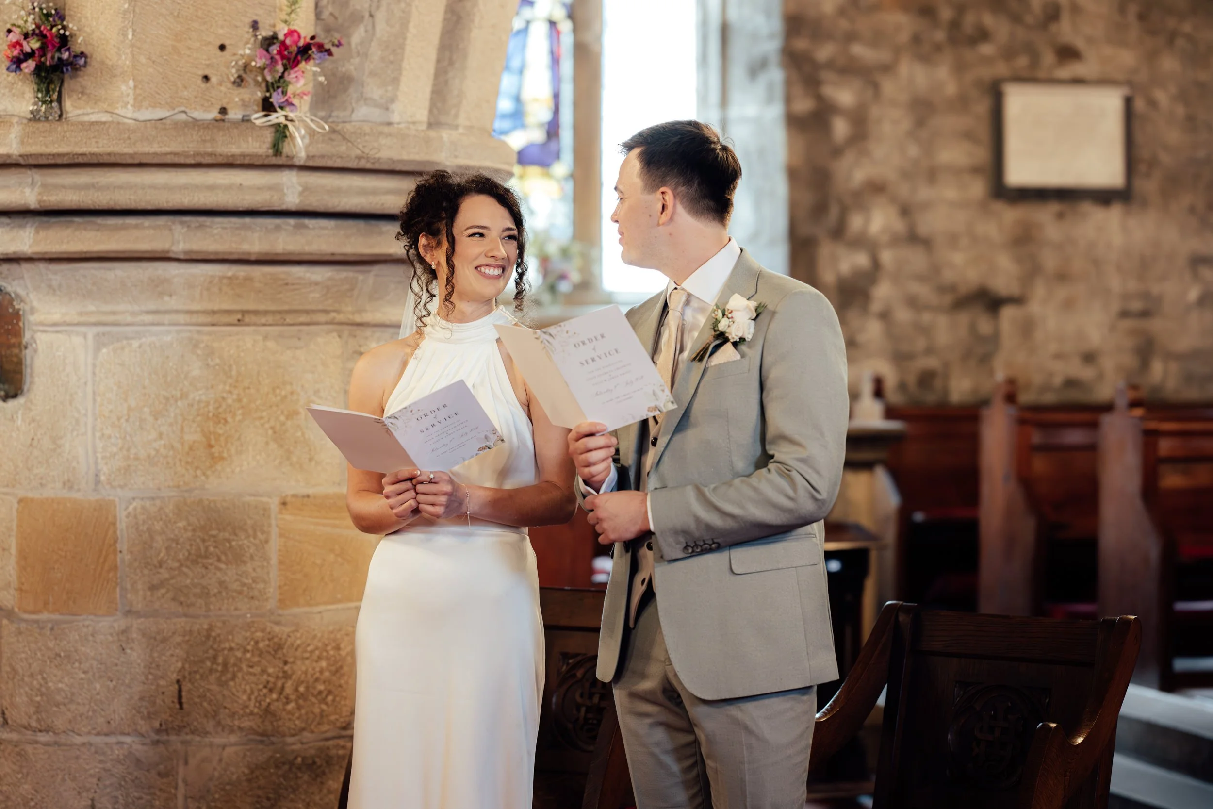 A bride and groom exchanging vows and smiling at each other during their wedding ceremony inside a church. The bride is wearing a white dress and holding a program; the groom is in a light suit with a flower boutonniere. The background includes stone