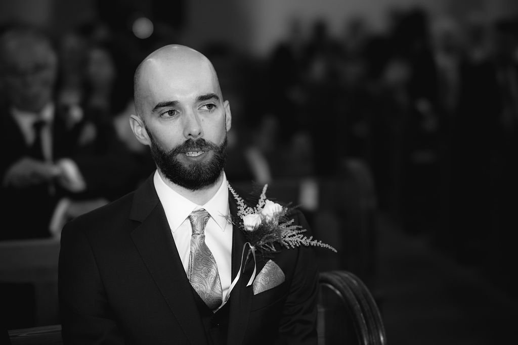 A man in a suit with a boutonnière, sitting in a formal setting, likely a wedding or ceremony, with several people blurred in the background.