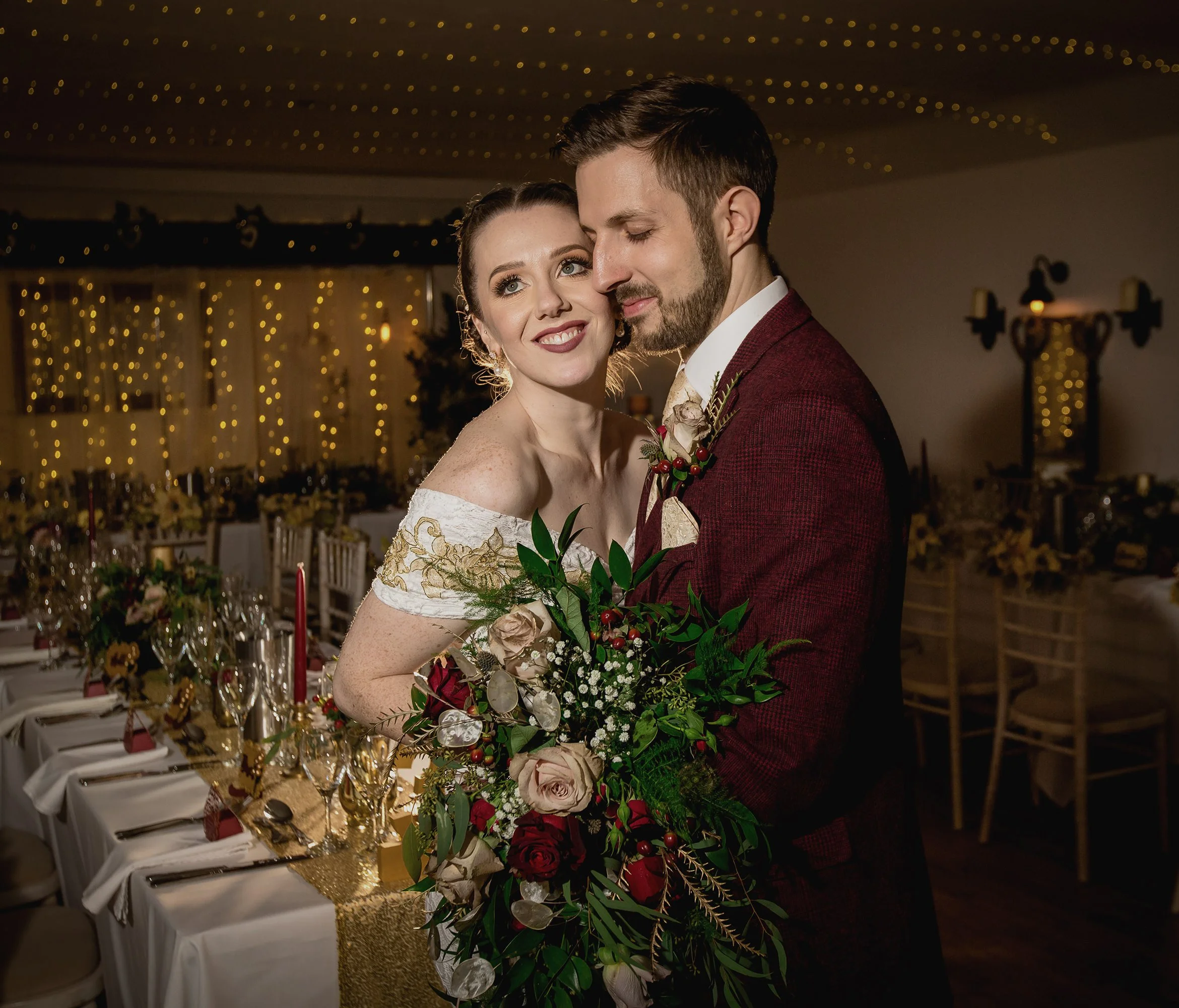 A bride and groom at their wedding reception, standing close together with the bride holding a large bouquet of flowers. The reception hall is decorated with string lights and set with elegant tables.
