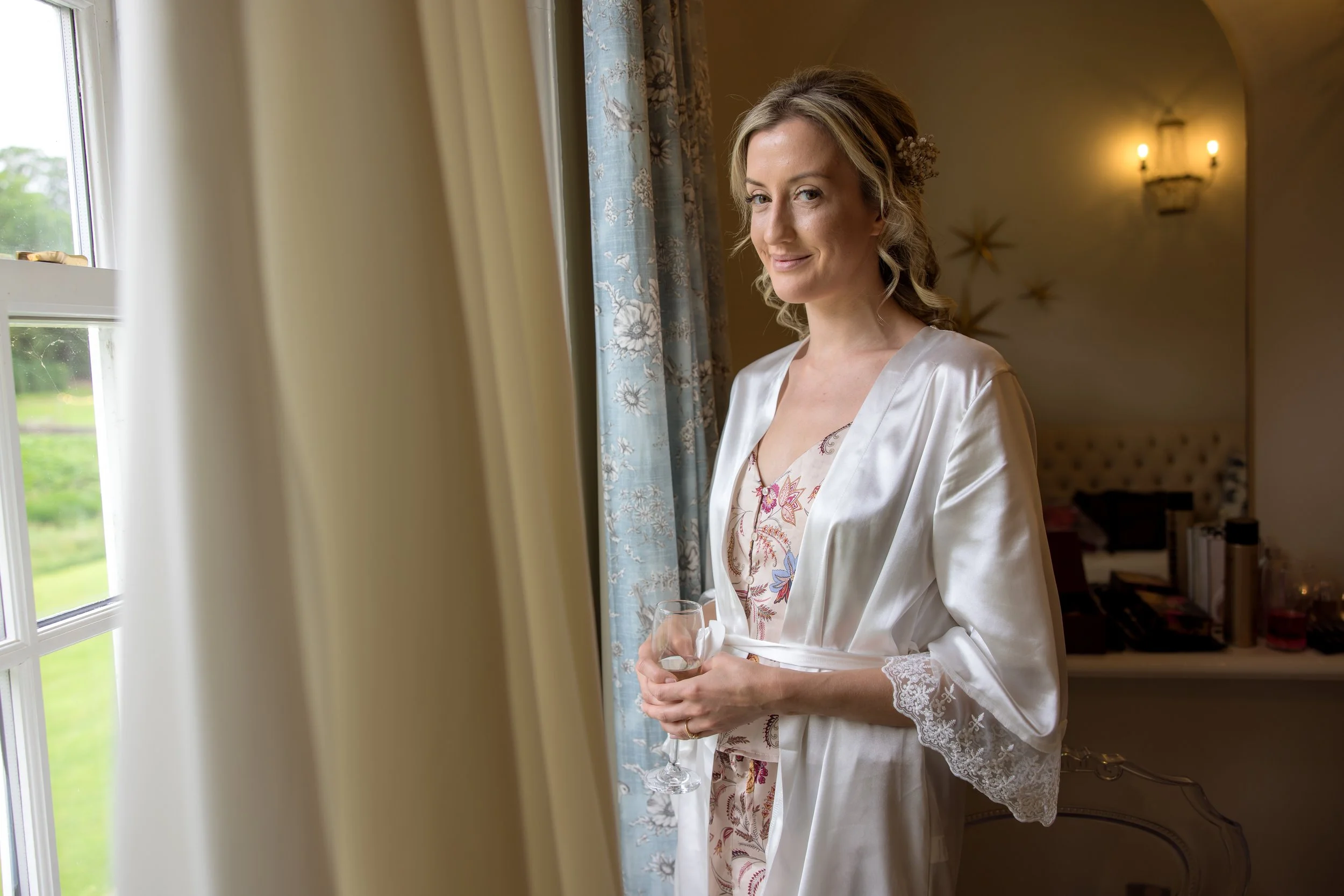 A woman in a satin robe holding a glass of wine, standing near a window, smiling softly, with a warmly lit room in the background.
