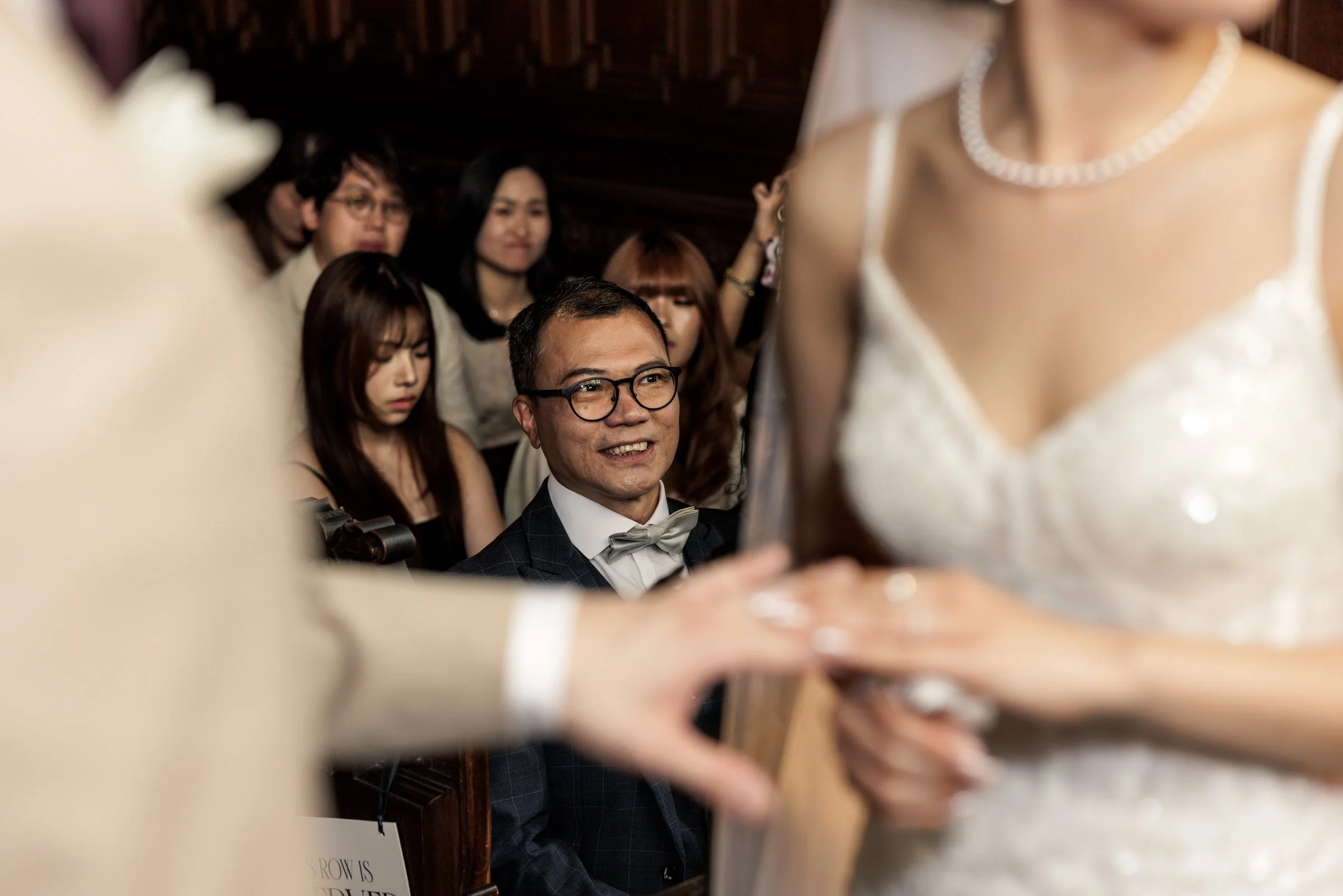 A group of diverse guests at a wedding reception, seated and smiling, with a focus on a man with glasses and a bow tie, viewed through a narrow gap in a decorative wooden structure. The bride wearing a white wedding dress and pearl necklace is partia