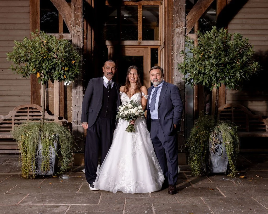 A bride in a white wedding dress holding a bouquet, flanked by two men in suits, standing in front of a wooden building with potted plants and trees.