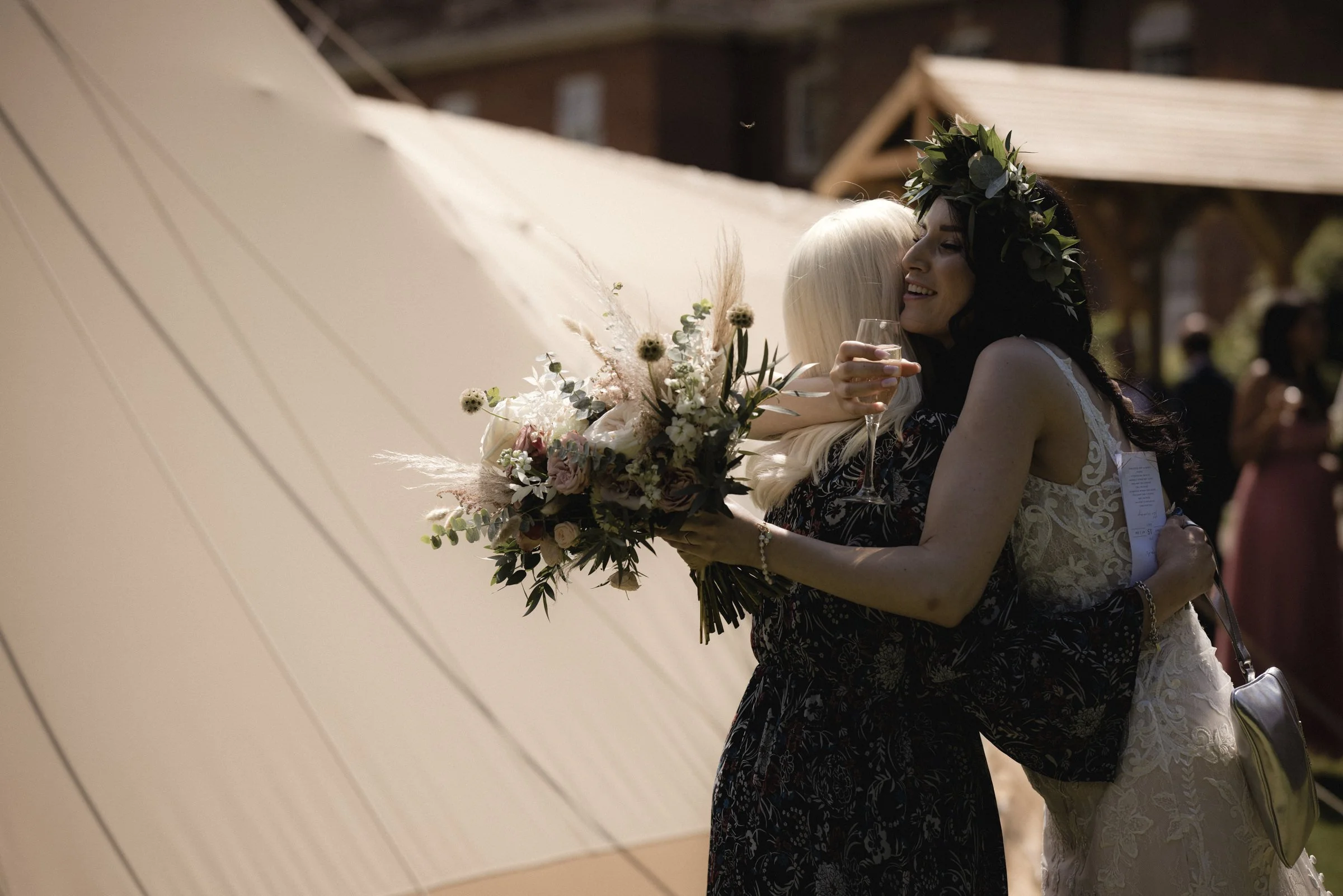 Two women hugging at a wedding, one in a white lace dress with a floral crown and the other holding a bouquet of flowers, with one holding a glass of champagne, in an outdoor setting with wooden structures.