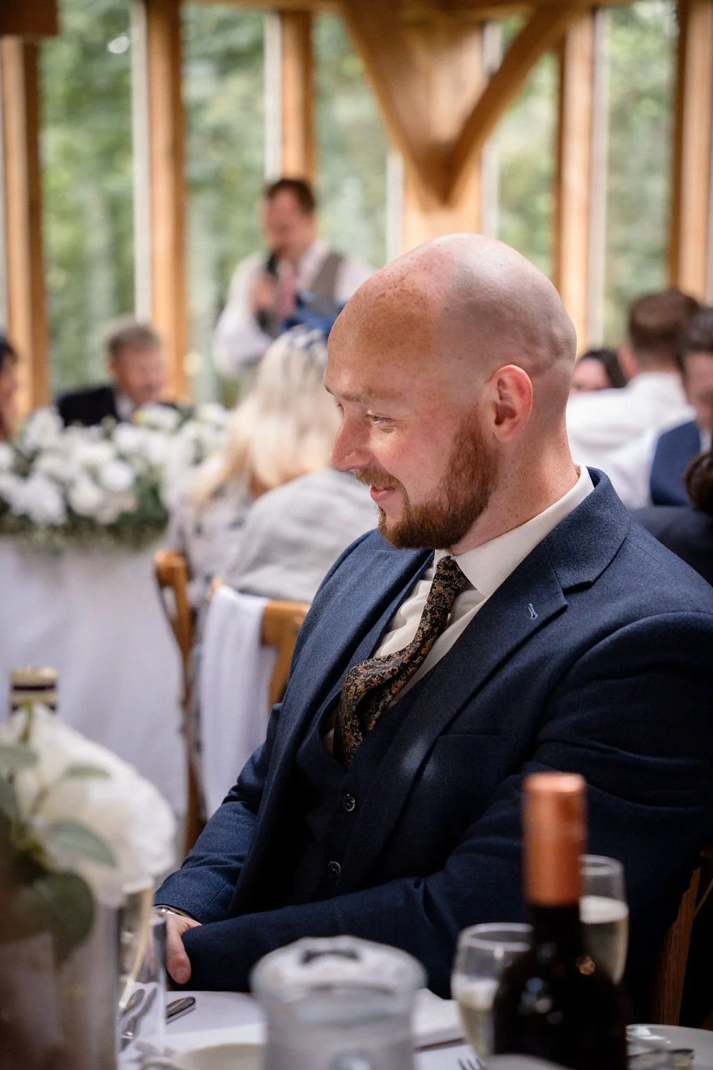 A man with a bald head and beard wearing a navy blue suit, sitting at a table during a wedding reception, smiling and looking down.