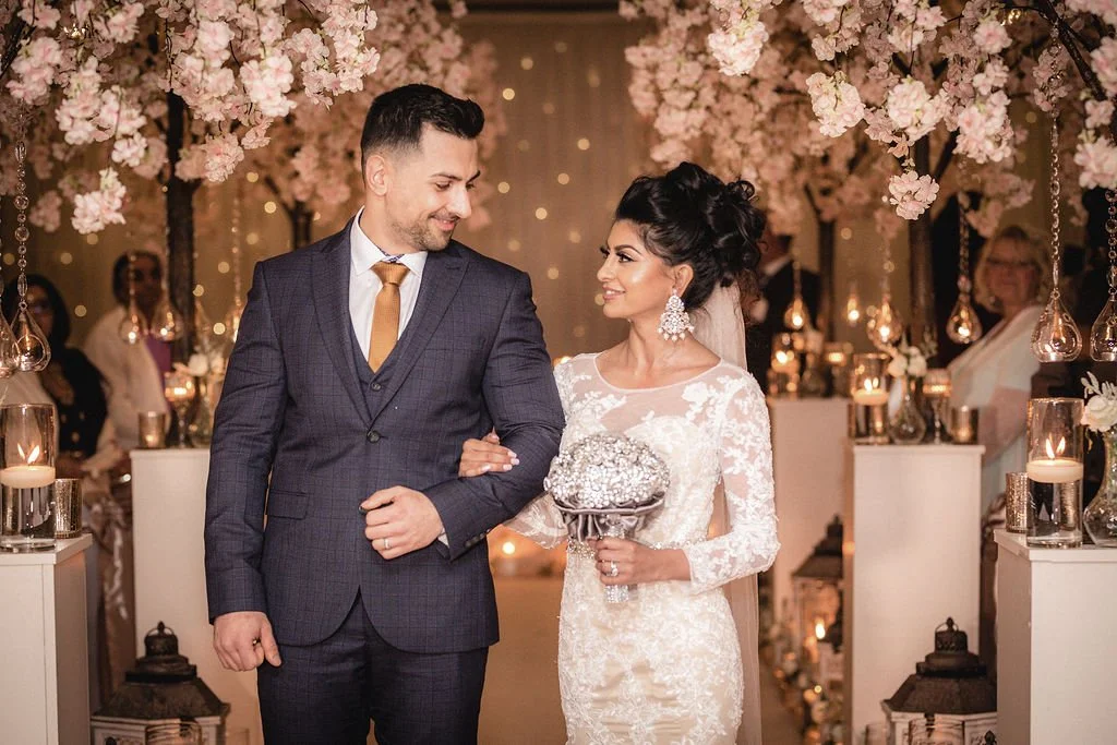 A bride and groom smiling at each other during their wedding ceremony, surrounded by pink flowers and decorative candles.