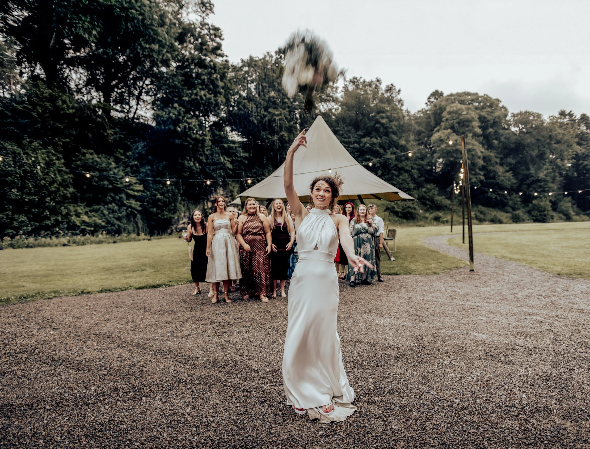 A woman in a white dress throws a bouquet into the air at an outdoor wedding reception, with a group of guests smiling behind her. Tents and string lights are visible in the background.