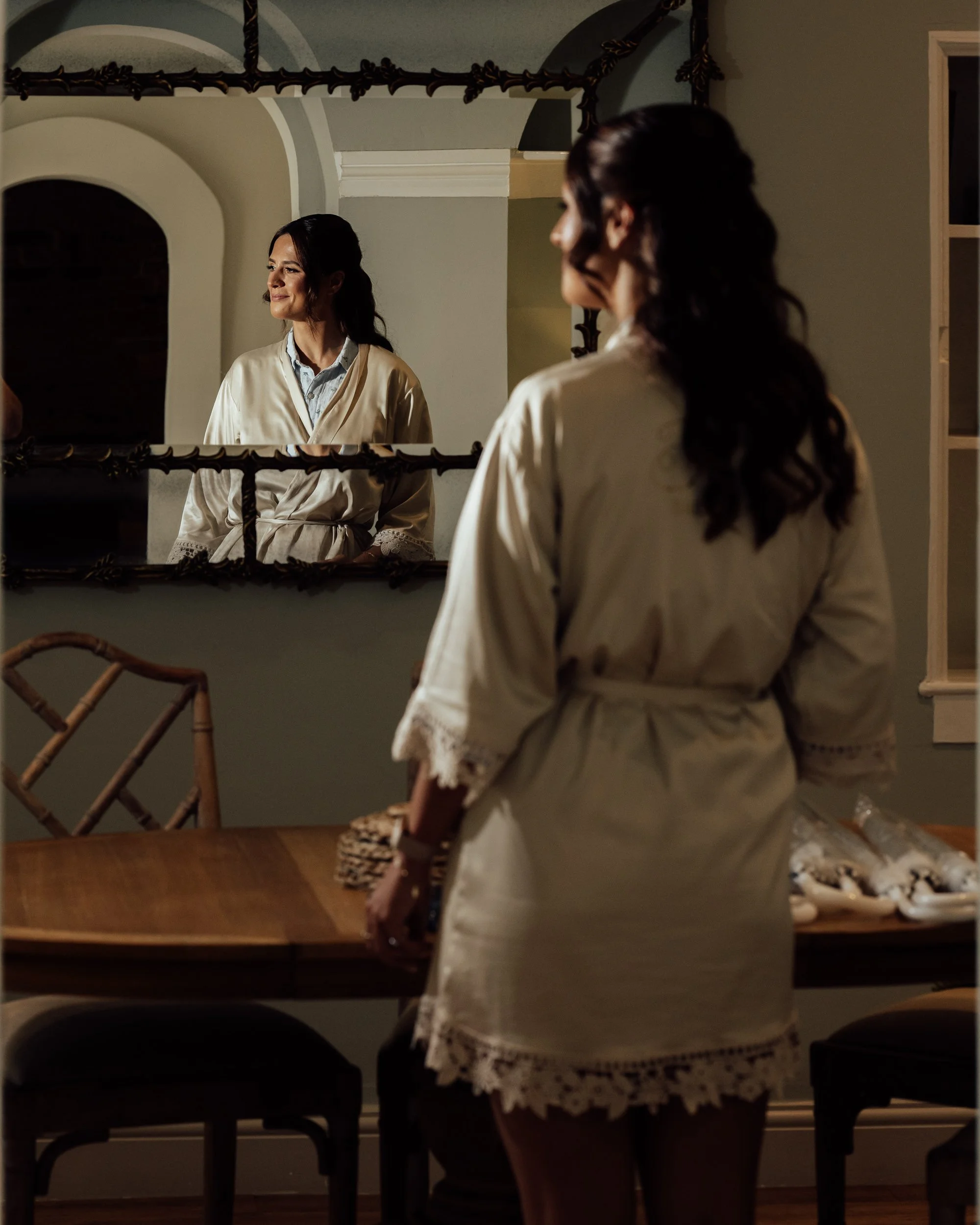 A woman looking at herself in a mirror, wearing a light-colored silk robe with lace detailing, standing in a dining room with a wooden table and chairs