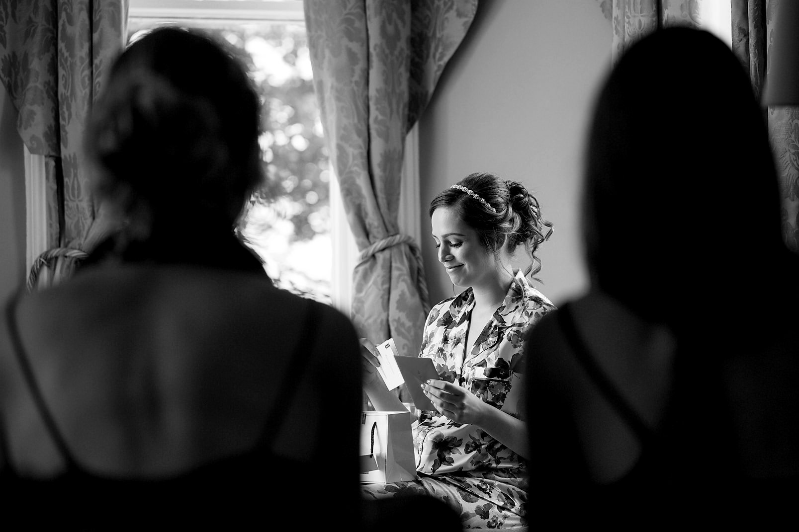 Black and white photo of a woman in a floral robe reading a card, with two blurred women in the foreground, inside a room with floral curtains and a window.