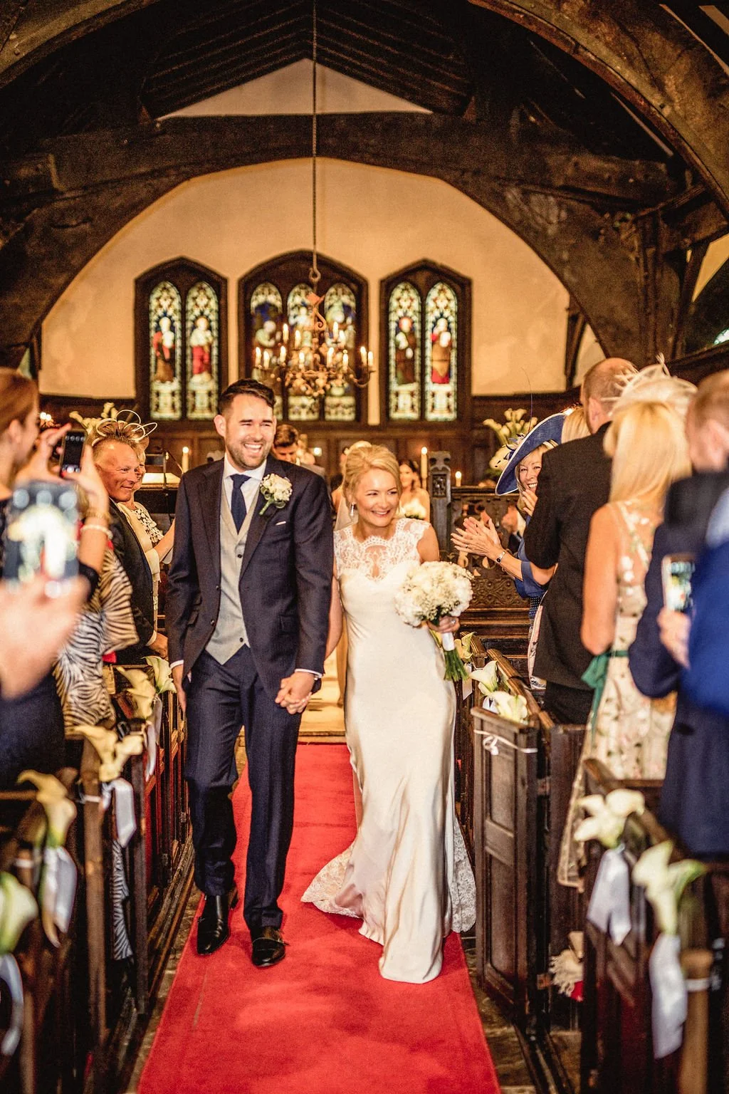A newlywed couple walking down the aisle inside a church, surrounded by smiling guests clapping and taking photos, with stained glass windows and a chandelier in the background.