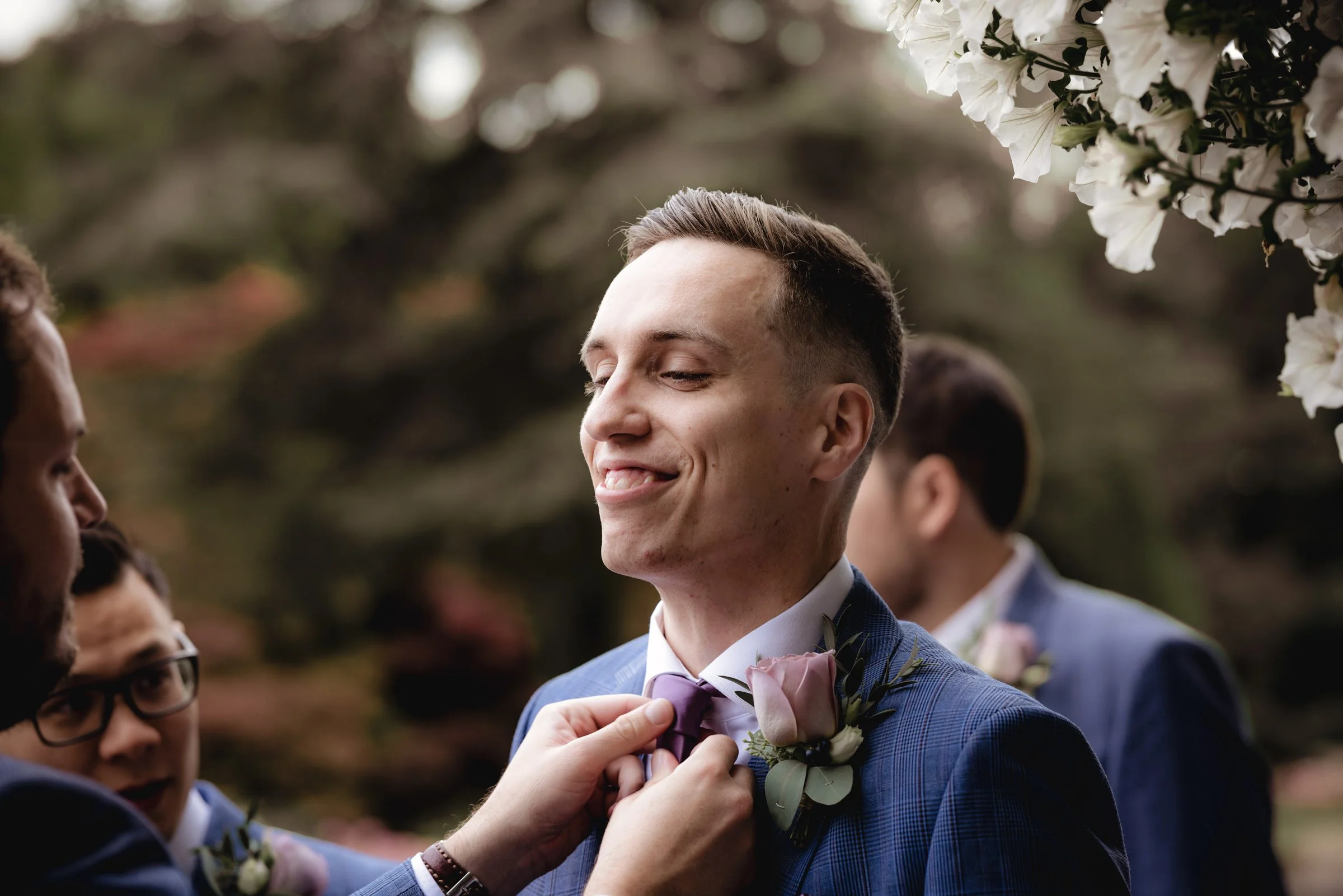 Man in blue suit with purple bow tie and boutonniere smiling with eyes closed during wedding ceremony, with another man adjusting his tie, outside with trees and white flowers in background.