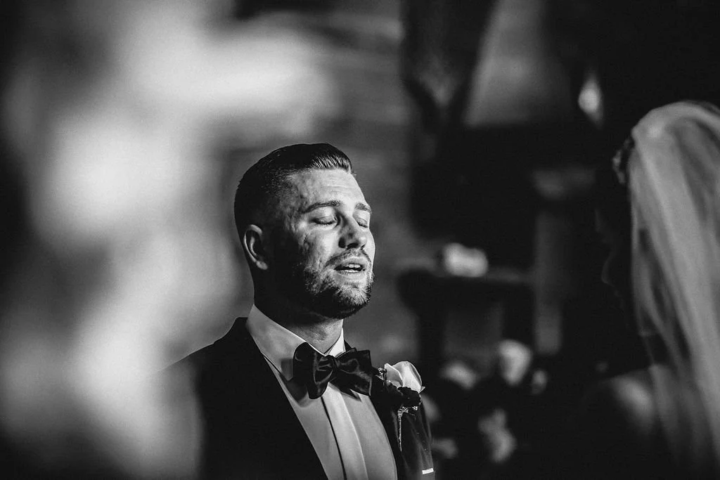 A man in a tuxedo with a bow tie, eyes closed, appears to be experiencing an emotional moment, possibly at a wedding, with a woman in a veil partially visible on the right.
