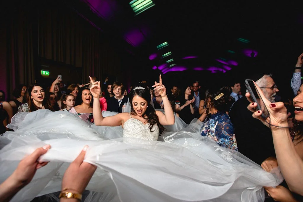 A bride dancing at her wedding reception surrounded by guests holding her wedding dress and taking photos.