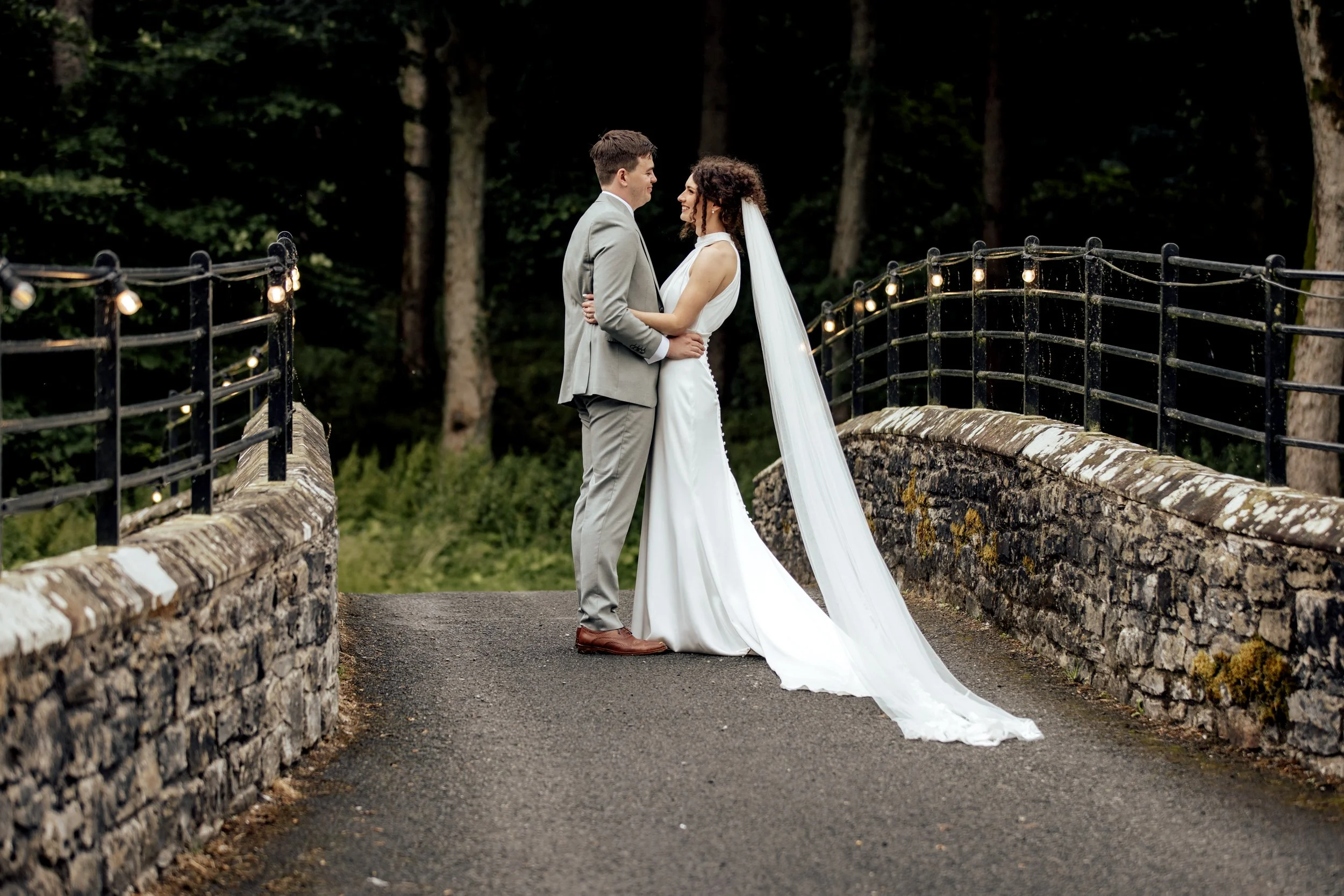 A bride and groom standing face-to-face on a small stone bridge in a wooded area, with decorative string lights along the railing, during a wedding ceremony.