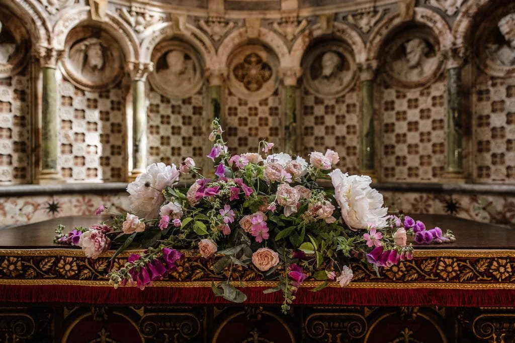 A floral arrangement with pink, purple, and white flowers on an ornate, decorated table in a grand, historic hall or church interior.