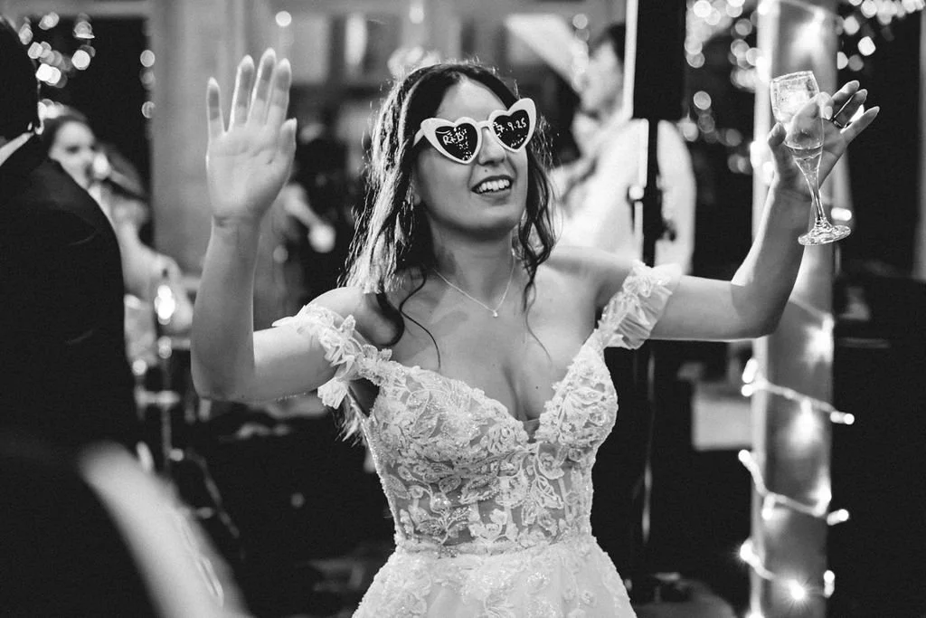 Woman celebrating at a party, wearing heart-shaped sunglasses, lace dress, and holding a wine glass, with balloons and fairy lights in the background.