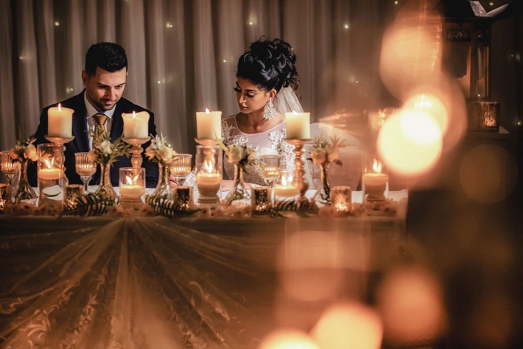 A bride and groom seated at a decorated wedding table with candles and flowers, surrounded by warm lighting.