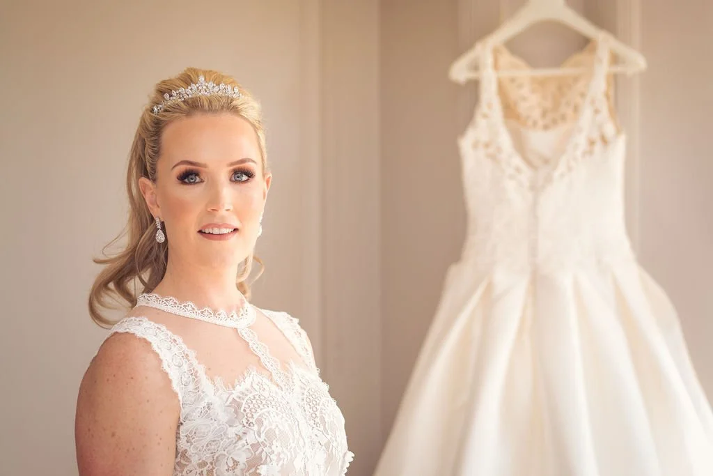 A bride in a lace wedding dress with a tiara and earrings, standing next to a hanging white wedding gown.