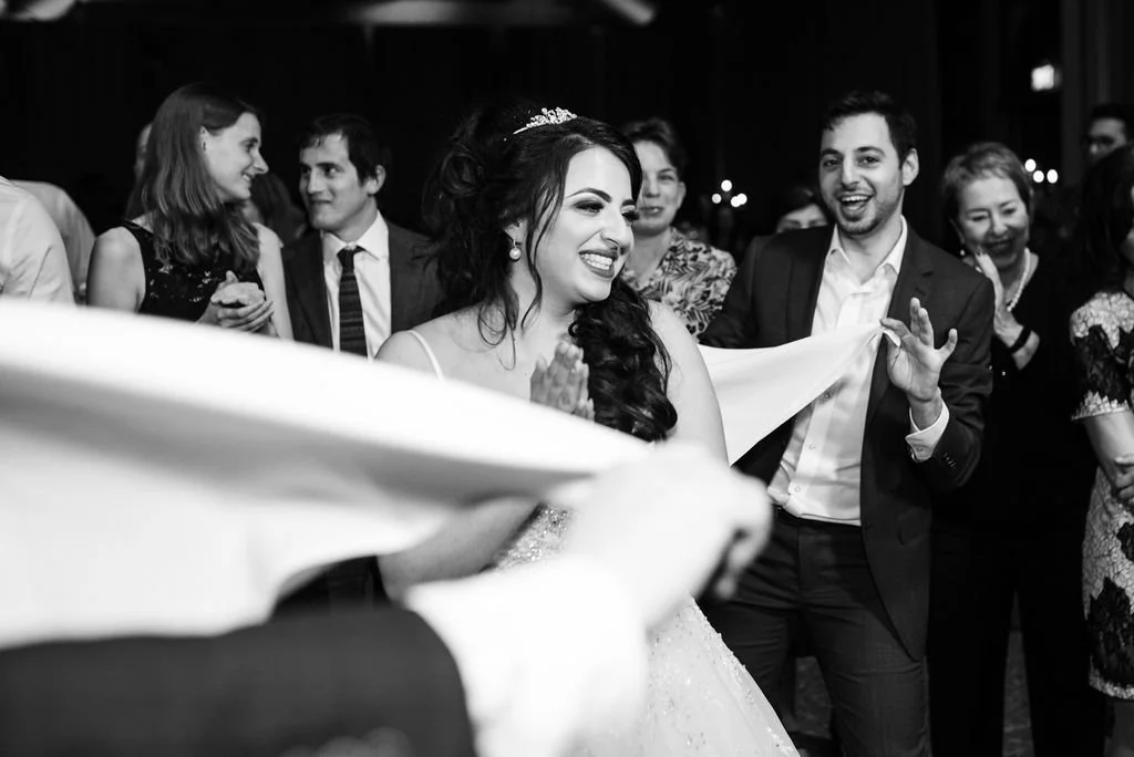 Black and white photo of a bride and groom dancing and laughing with guests at their wedding celebration.