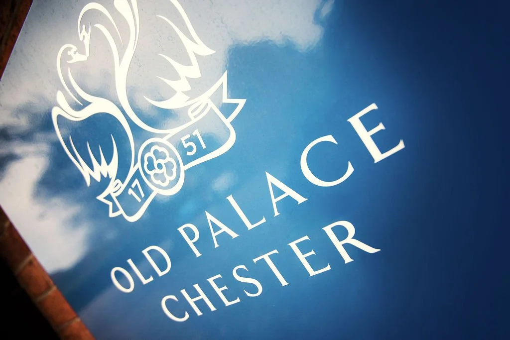 Close-up of a blue sign with white lettering that reads "Old Palace Chester" and an emblem with the year 1751 and a floral symbol.