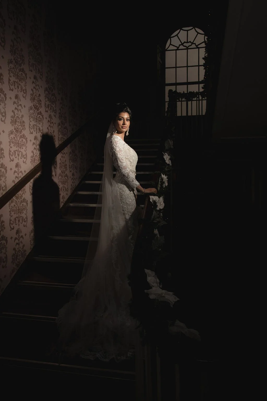 Bride in a white lace wedding dress standing beside a staircase with floral decorations, indoors near a window with a sunlit sky outside.
