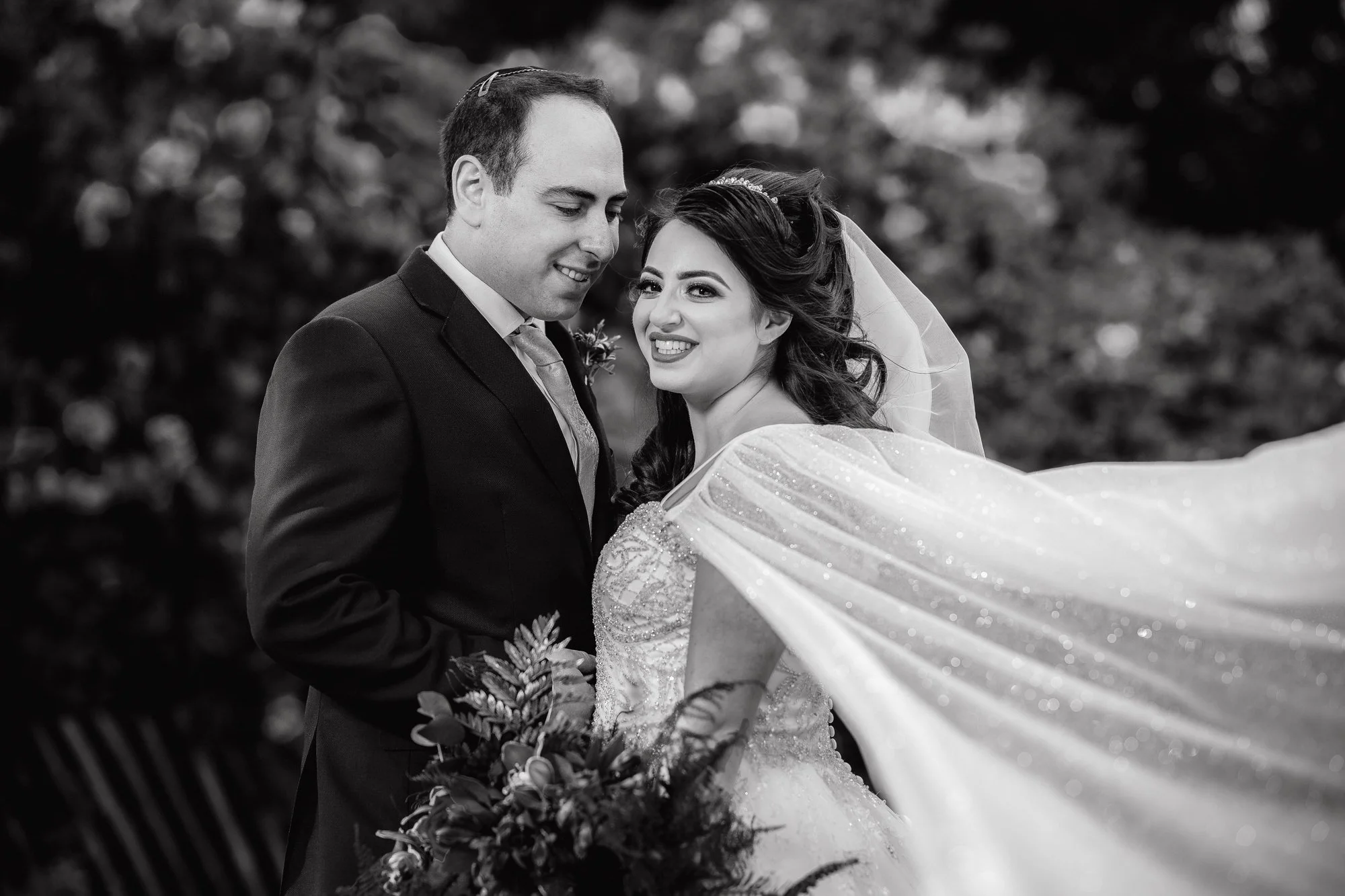 A black and white photo of a wedding couple outdoors, with the groom in a suit and the bride in a wedding dress, smiling and embracing each other.