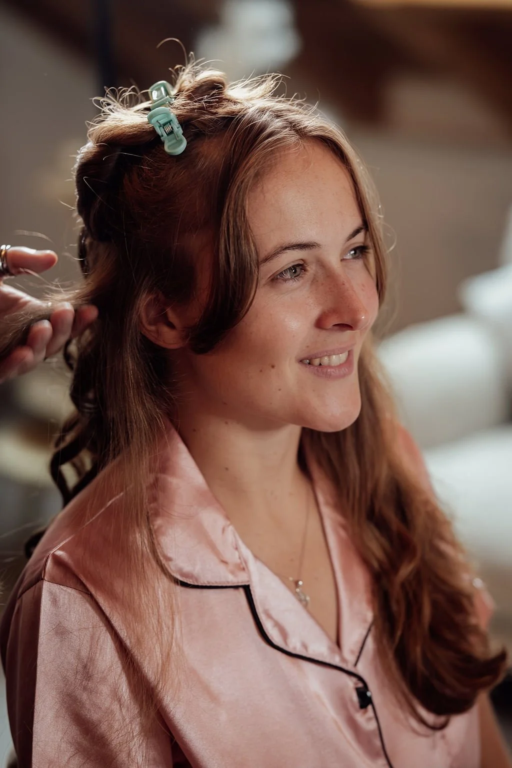 A woman with reddish hair getting her hair styled with curlers and clips, smiling as a hairstylist works on her hair.