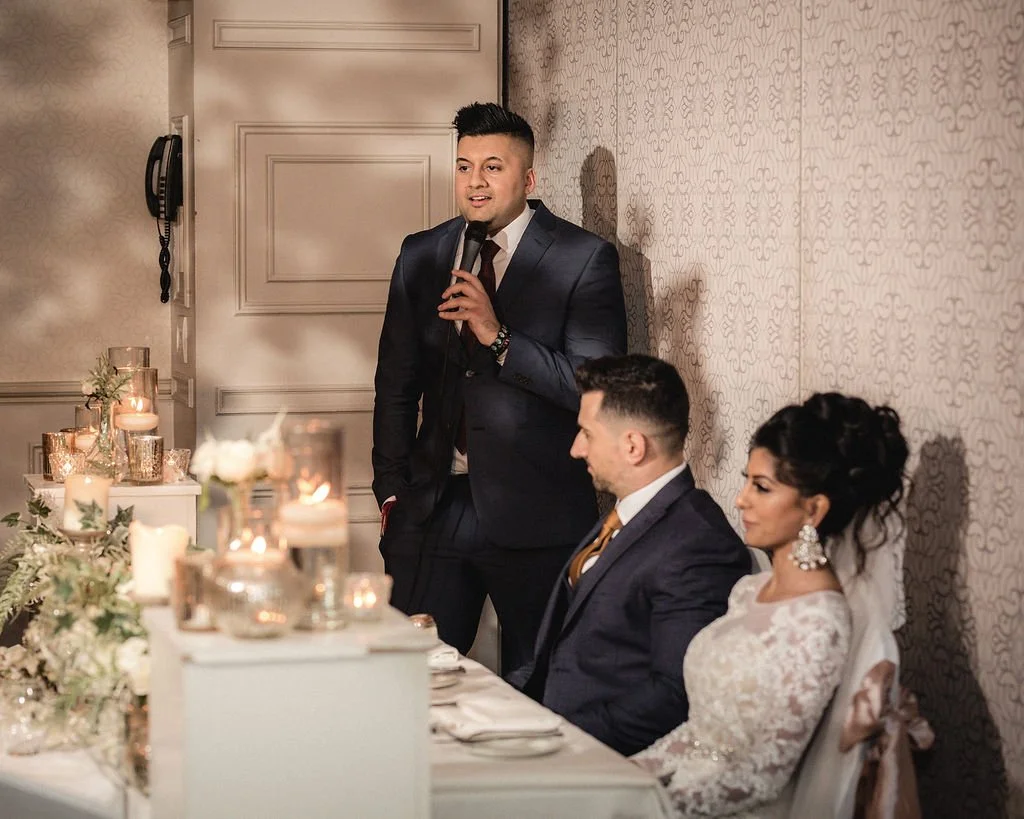 Man in a suit giving a speech at a wedding reception with the bride and groom seated at the table.