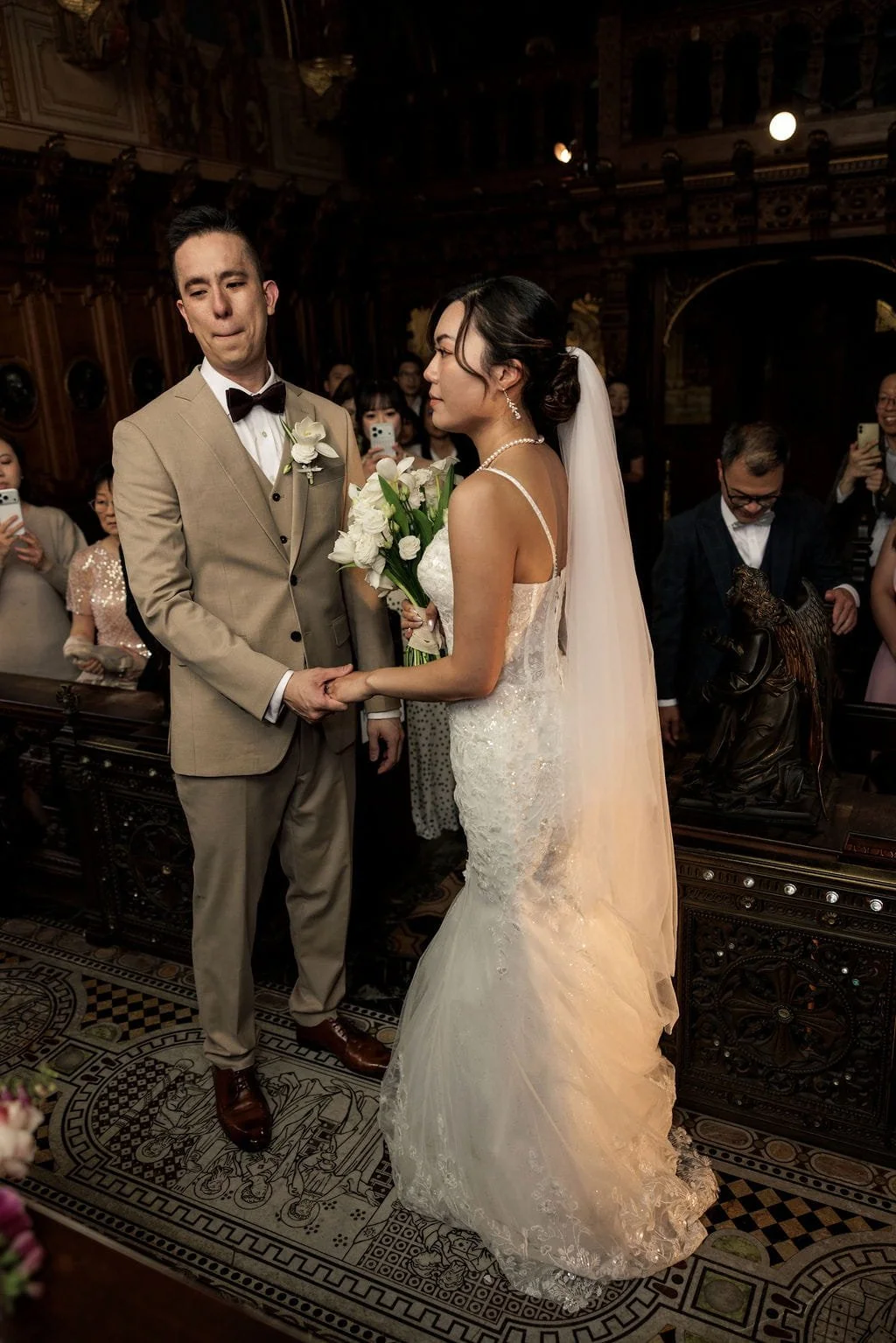 A bride and groom holding hands during their wedding ceremony in a decorated church, surrounded by guests taking photos.