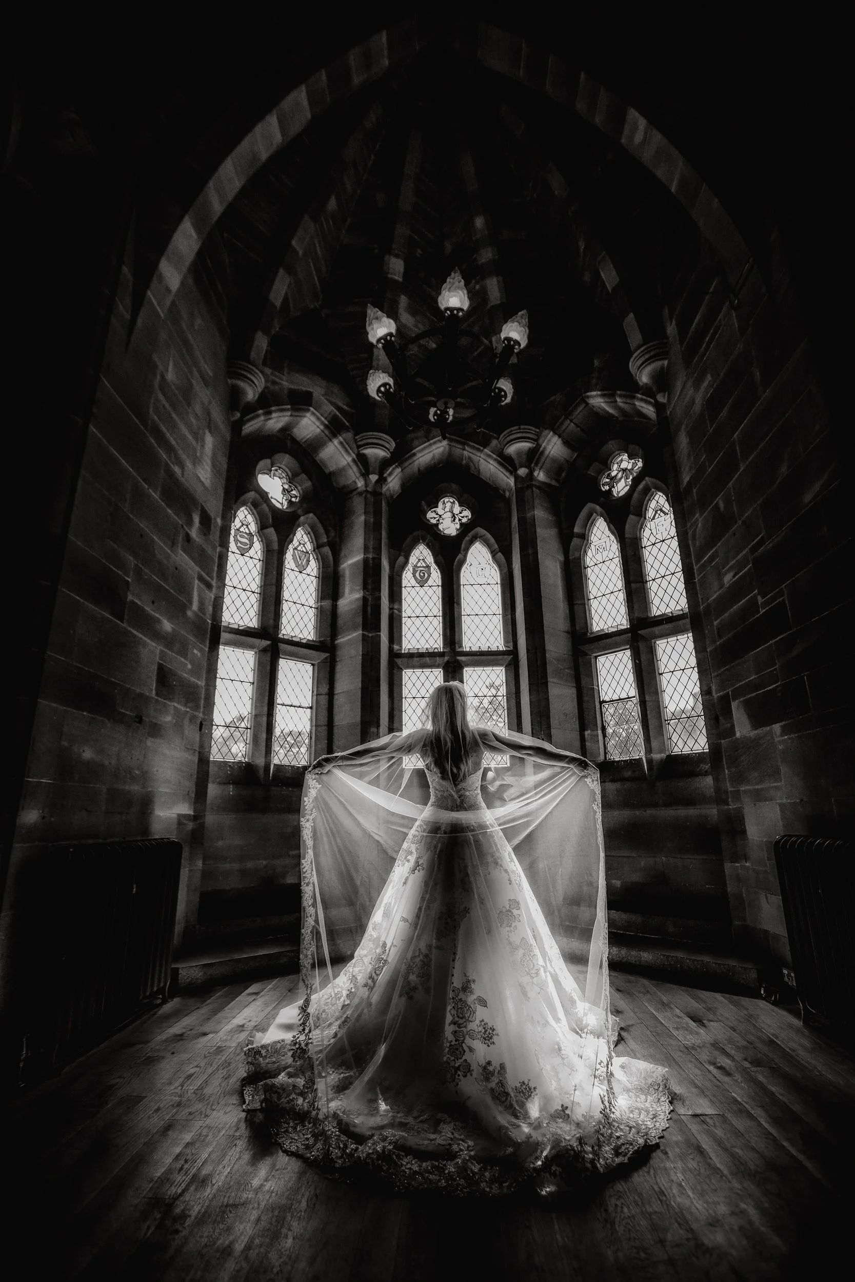 A woman in a wedding dress stands inside a gothic-style chapel with large stained glass windows, holding her veil outstretched behind her in a dramatic pose.