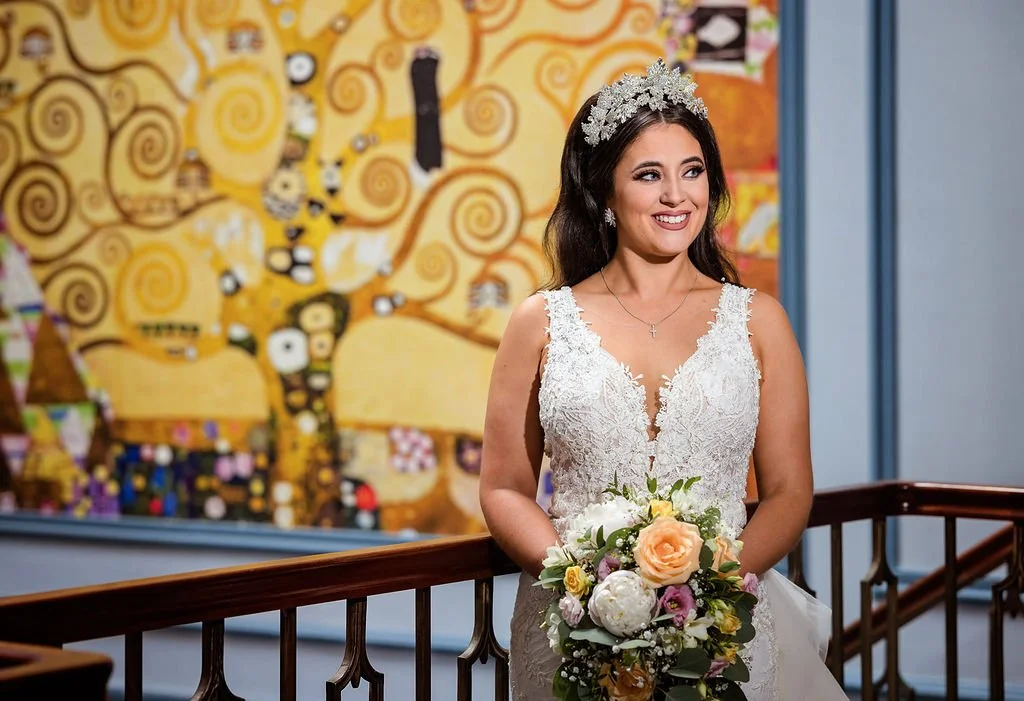 A bride in a white lace wedding dress holding a bouquet of flowers, standing indoors with a colorful abstract painting on the wall behind her.