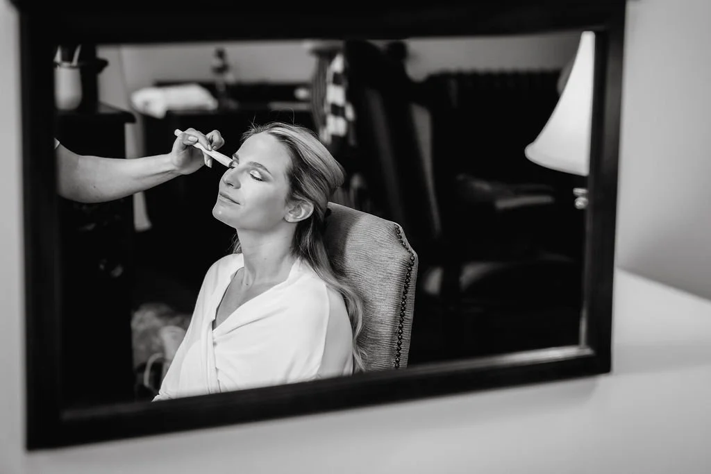 A woman getting her makeup done, reflected in a mirror, with her eyes closed and a peaceful expression.