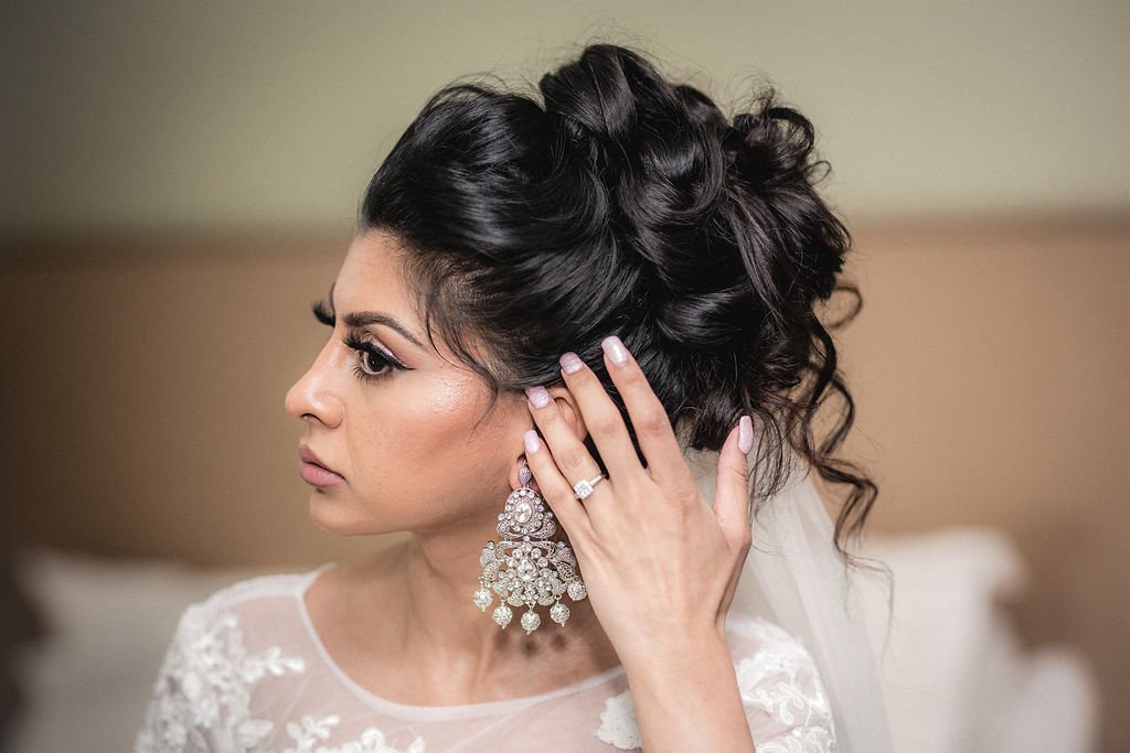 A bride with dark, styled hair, wearing elaborate earrings and a wedding gown, adjusting her earring while looking to the left.